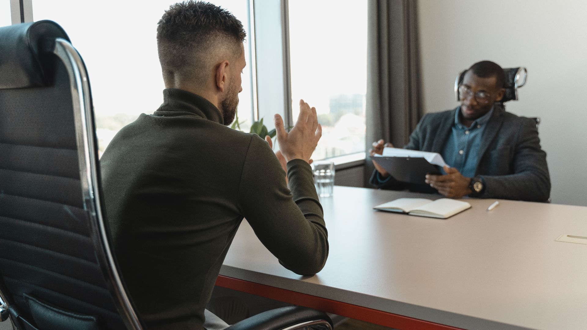Two men in a job interview setting in a modern office with large windows.