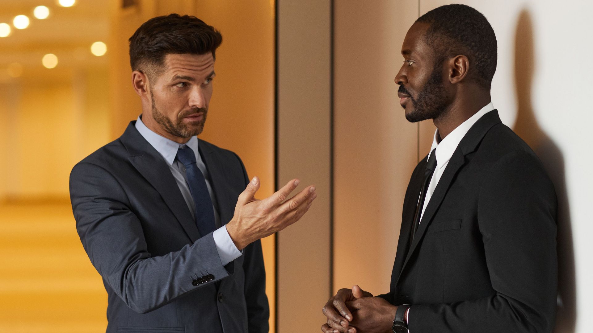 Two businessmen in formal attire engaged in a serious discussion in a modern office hallway.