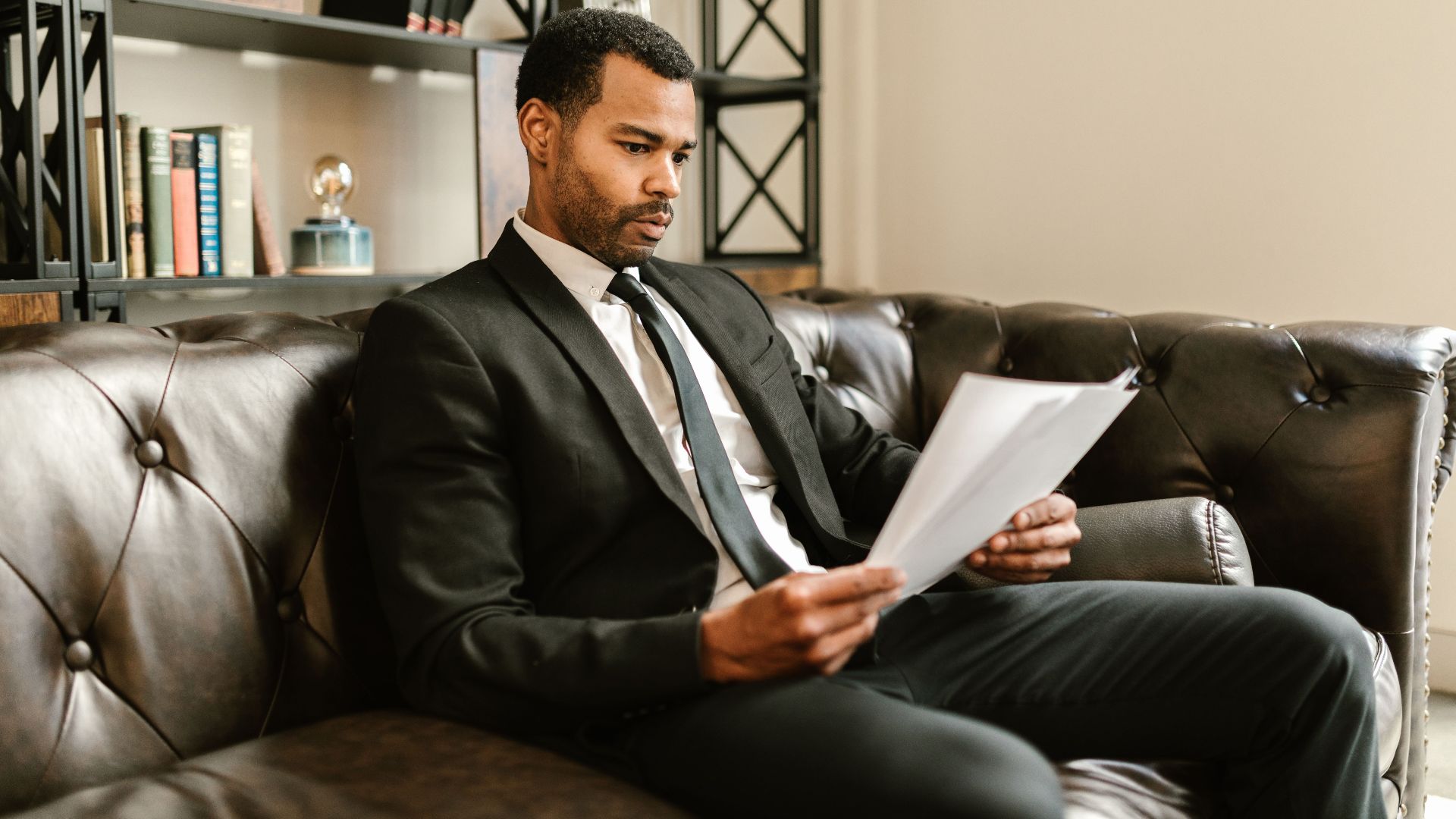 Businessman in suit reading papers on a leather sofa in a modern office setting.