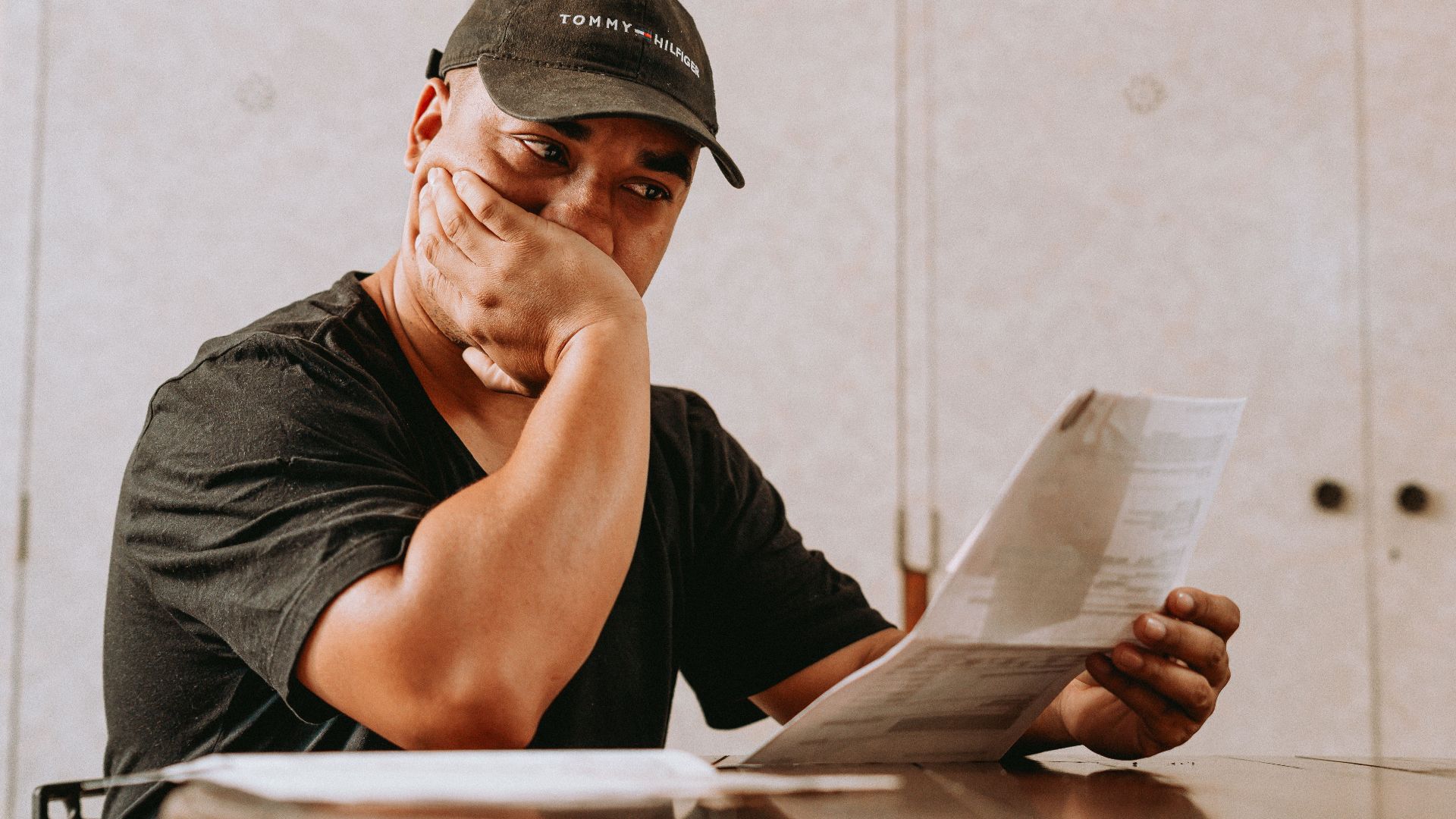 A man sits indoors, deeply focused on reviewing paperwork, expressing concern and contemplation.