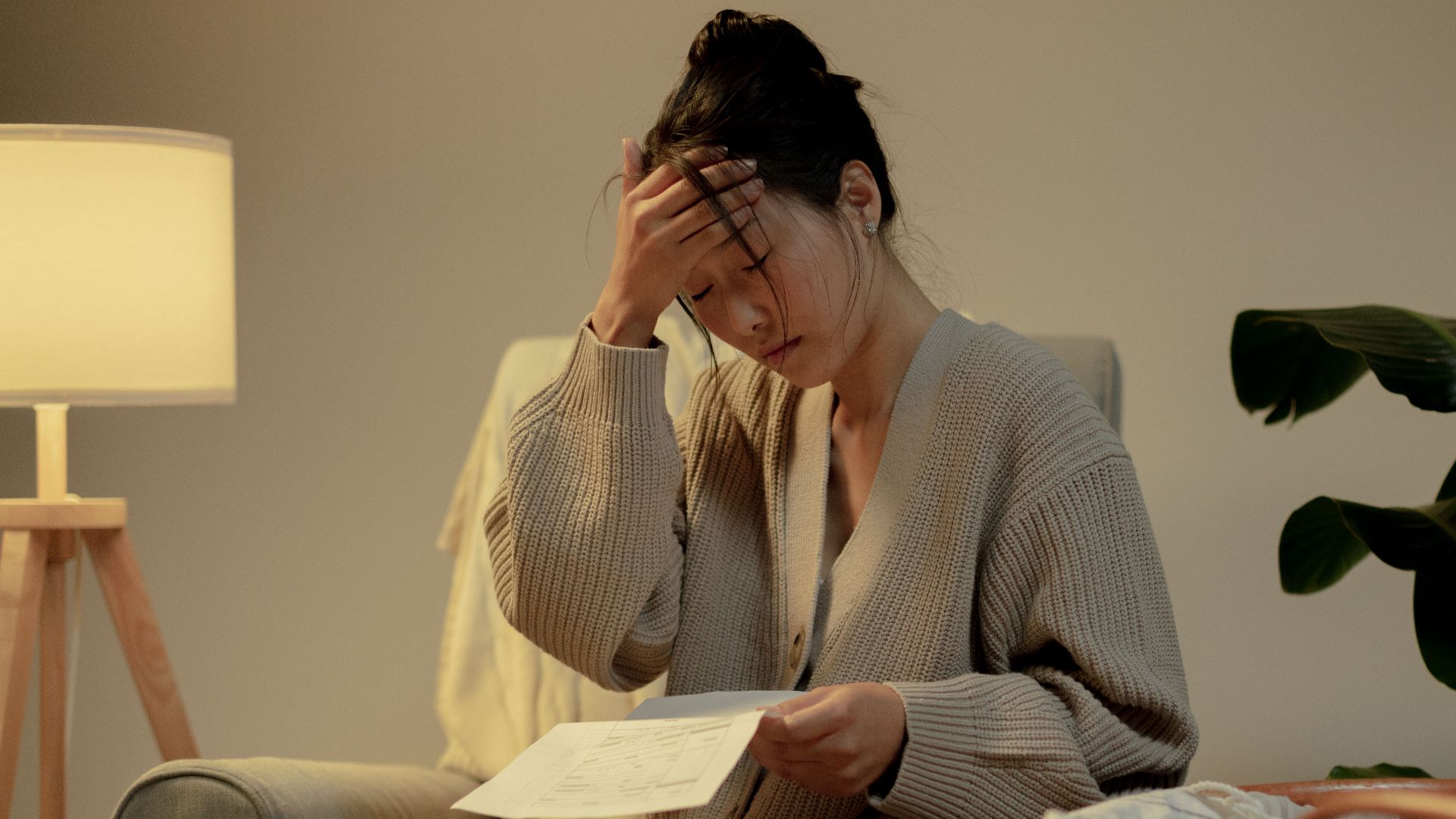A woman sits in a cozy room, stressed while reading paperwork, hand on head.