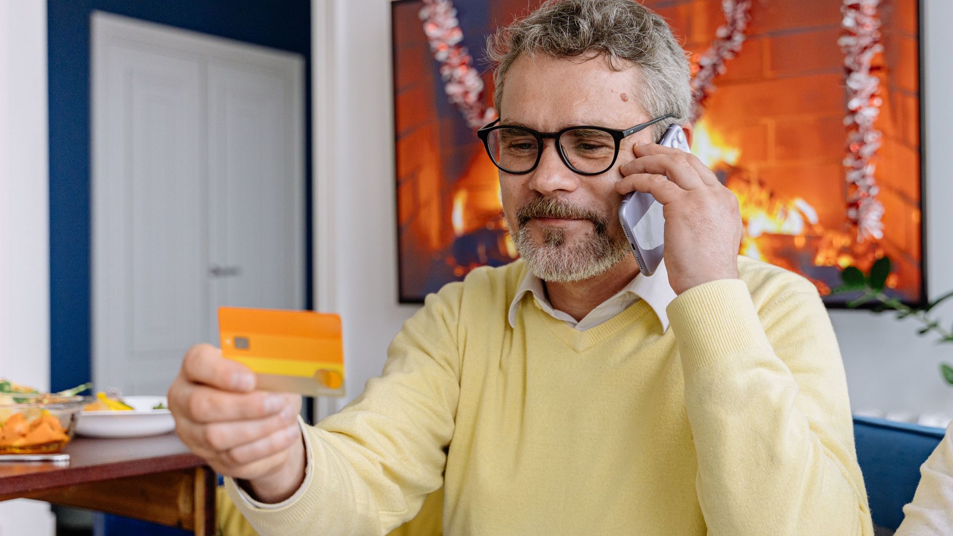 Elderly man using phone while holding a credit card indoors near a cozy fireplace.