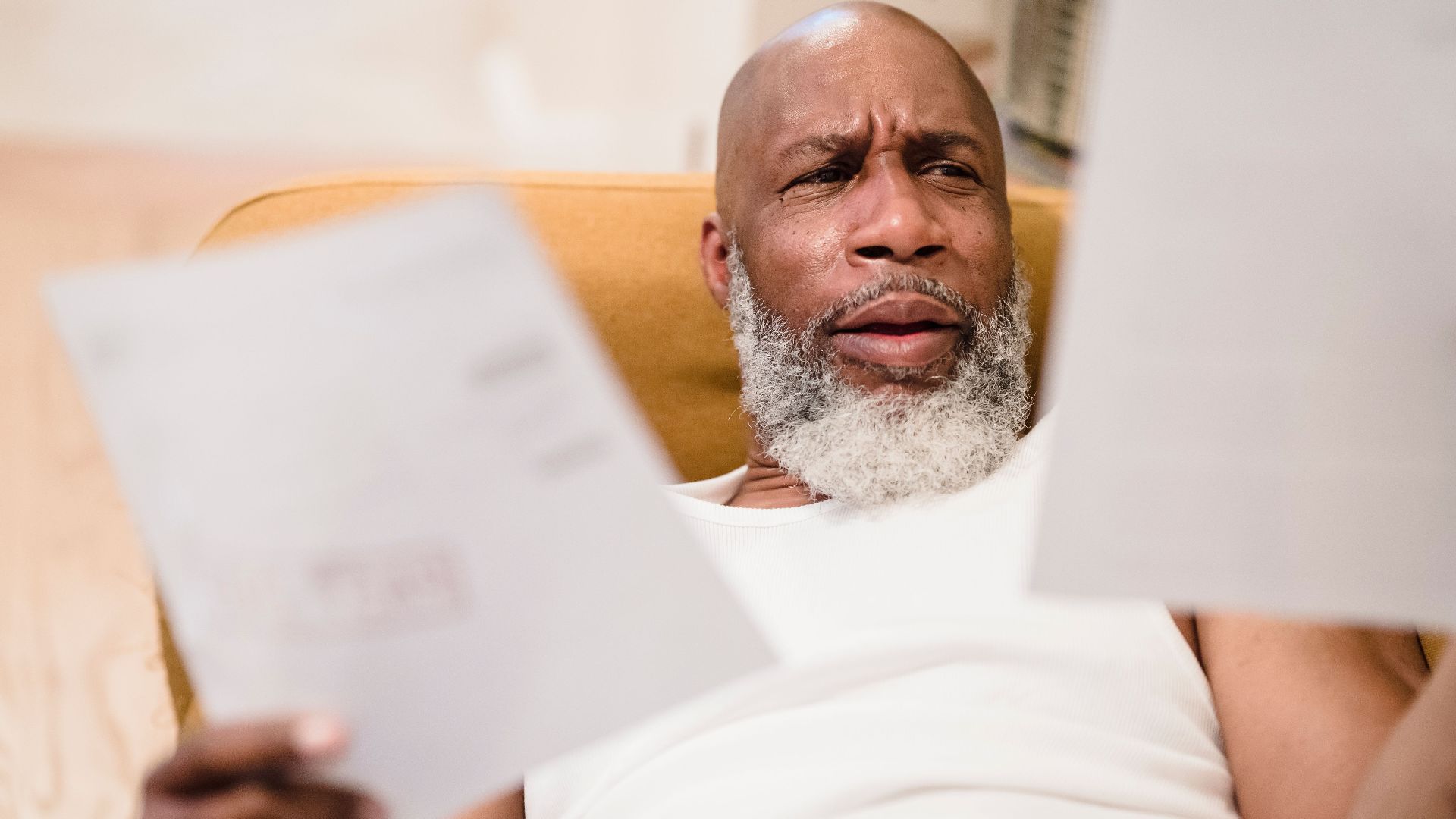 A middle-aged man looks concerned as he reviews household bills, sitting in a comfortable chair indoors.