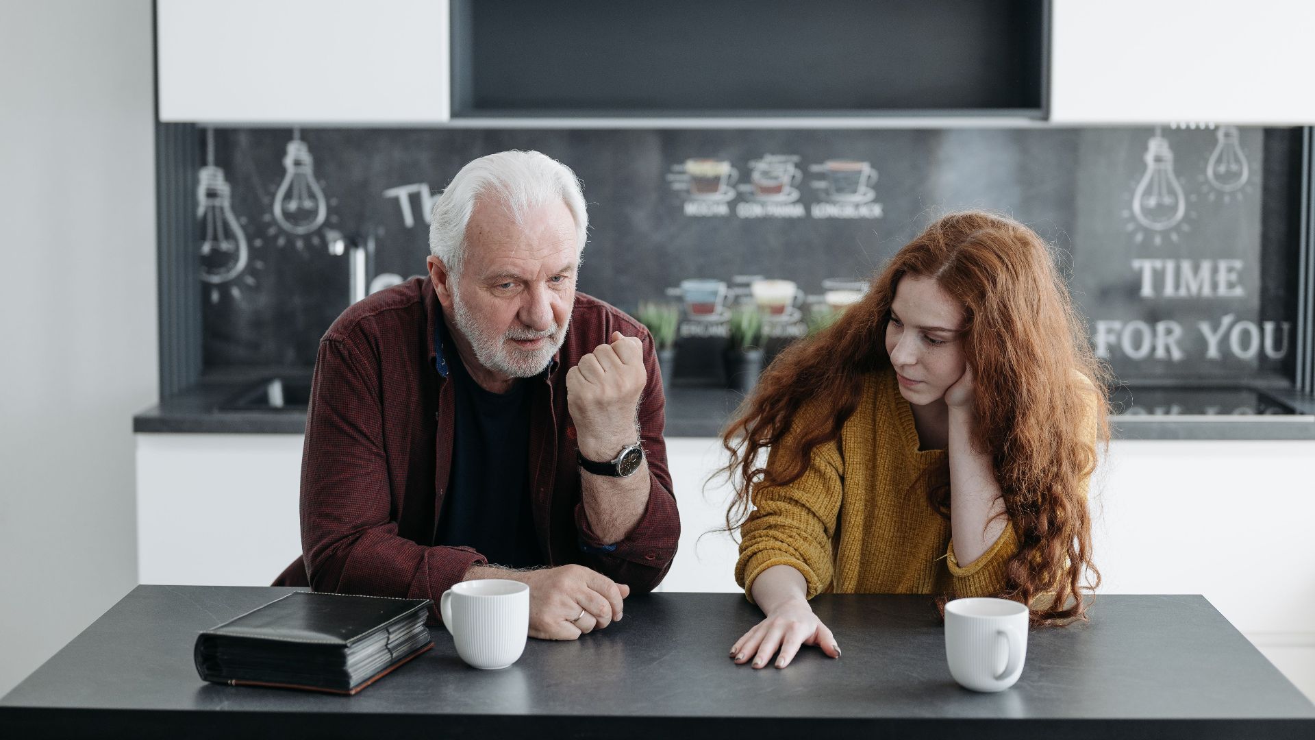 An elderly man and his granddaughter engage in a heartfelt conversation over coffee indoors.