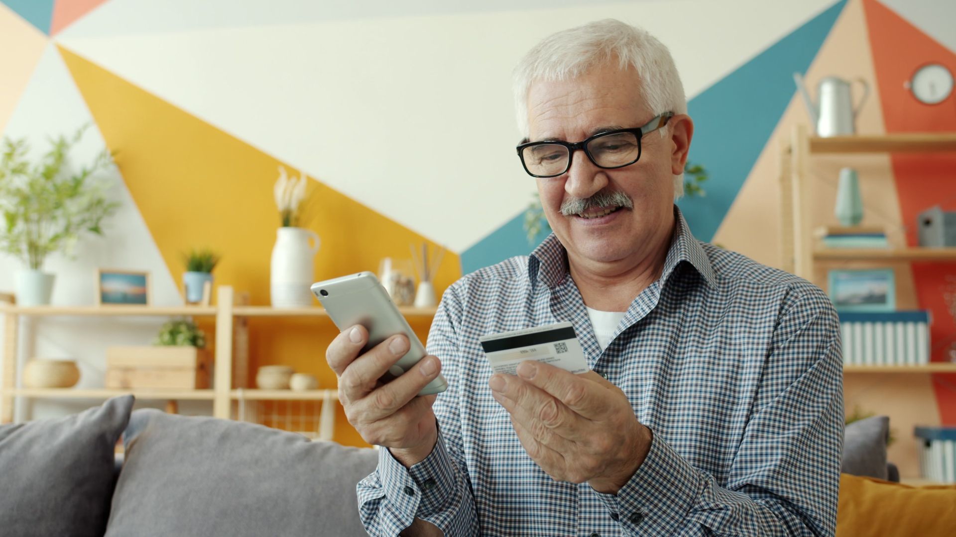 Senior man using smartphone and credit card for online shopping indoors.