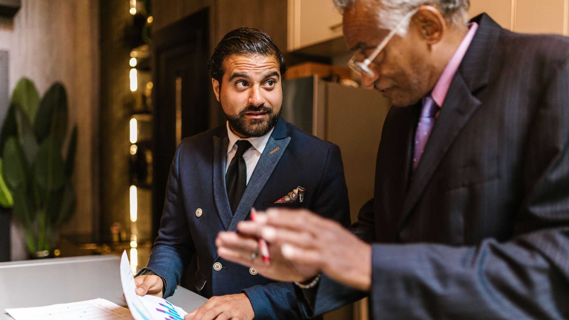 Two men in suits discuss financial documents with graphs indoors.