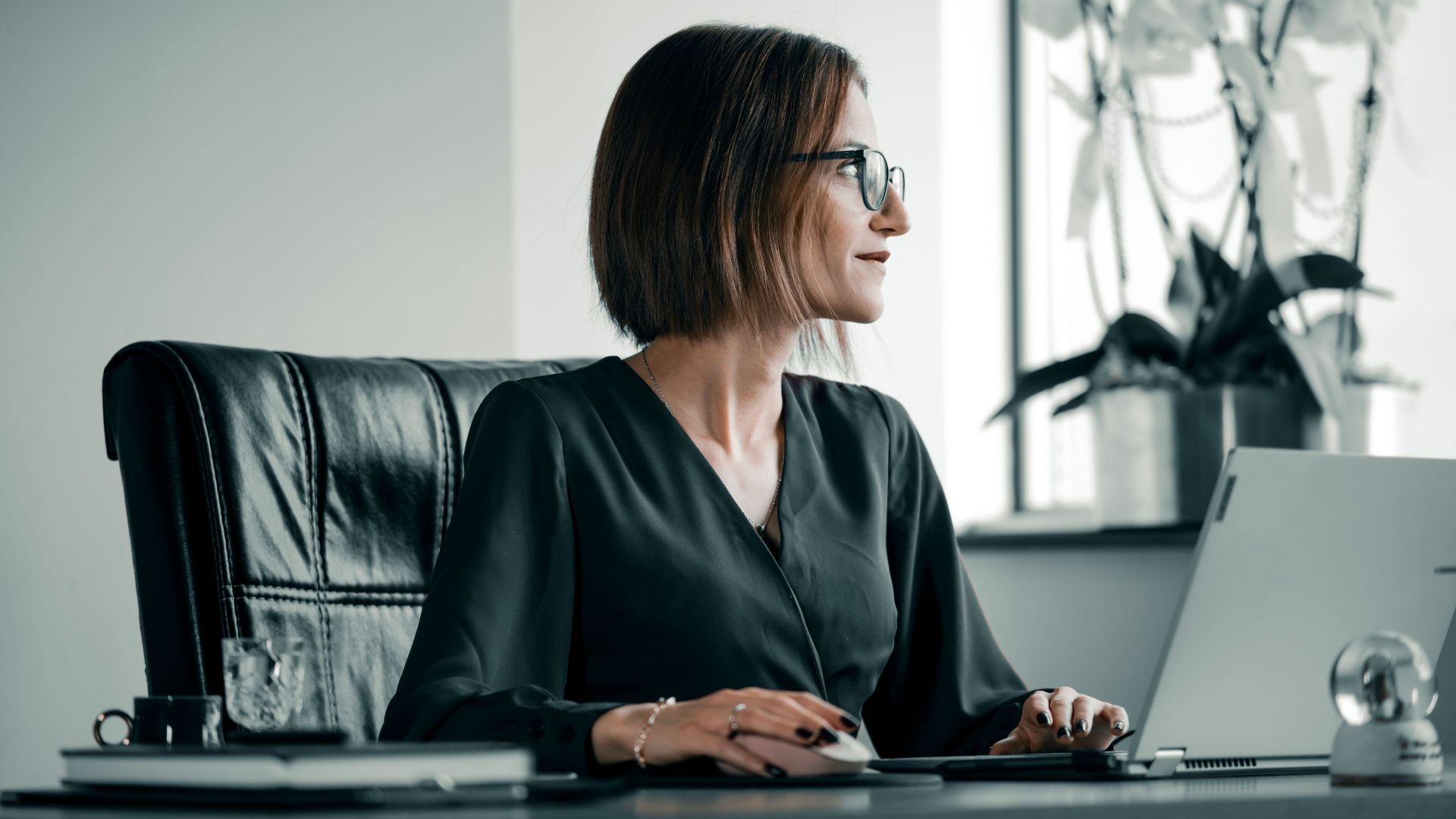 Adult woman working at office desk with laptop, glasses, and leather chair.