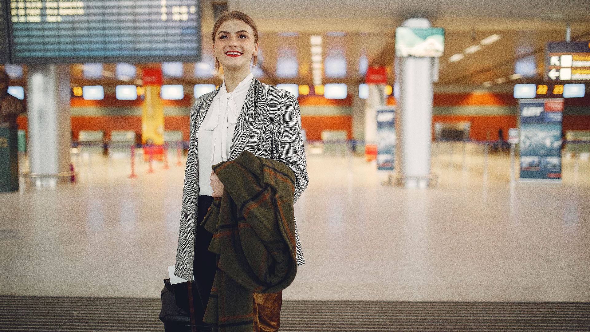 Smiling female manager in elegant wear checking schedule of flights in airport while standing with suitcase and coat looking up