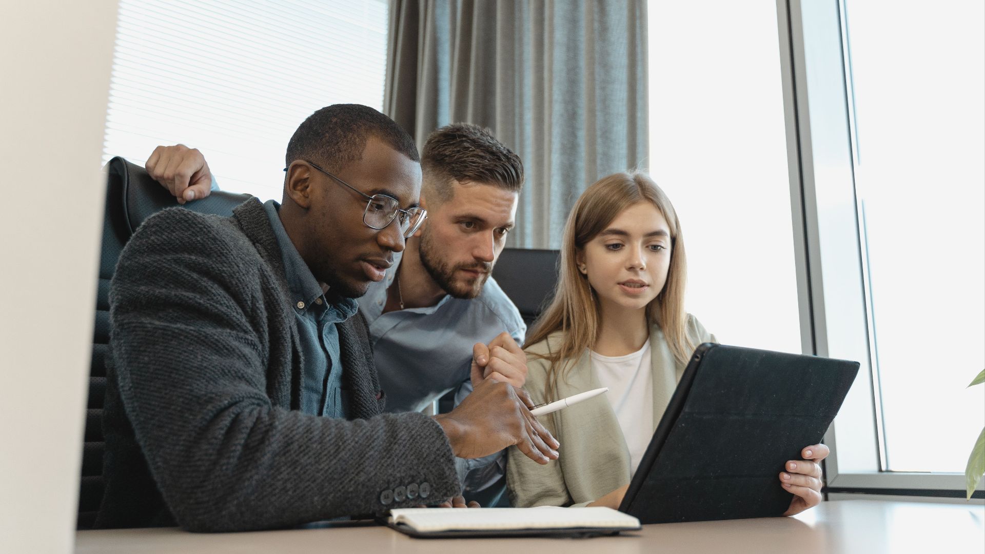 Team of three diverse professionals working on a project with a tablet in a modern office setting.