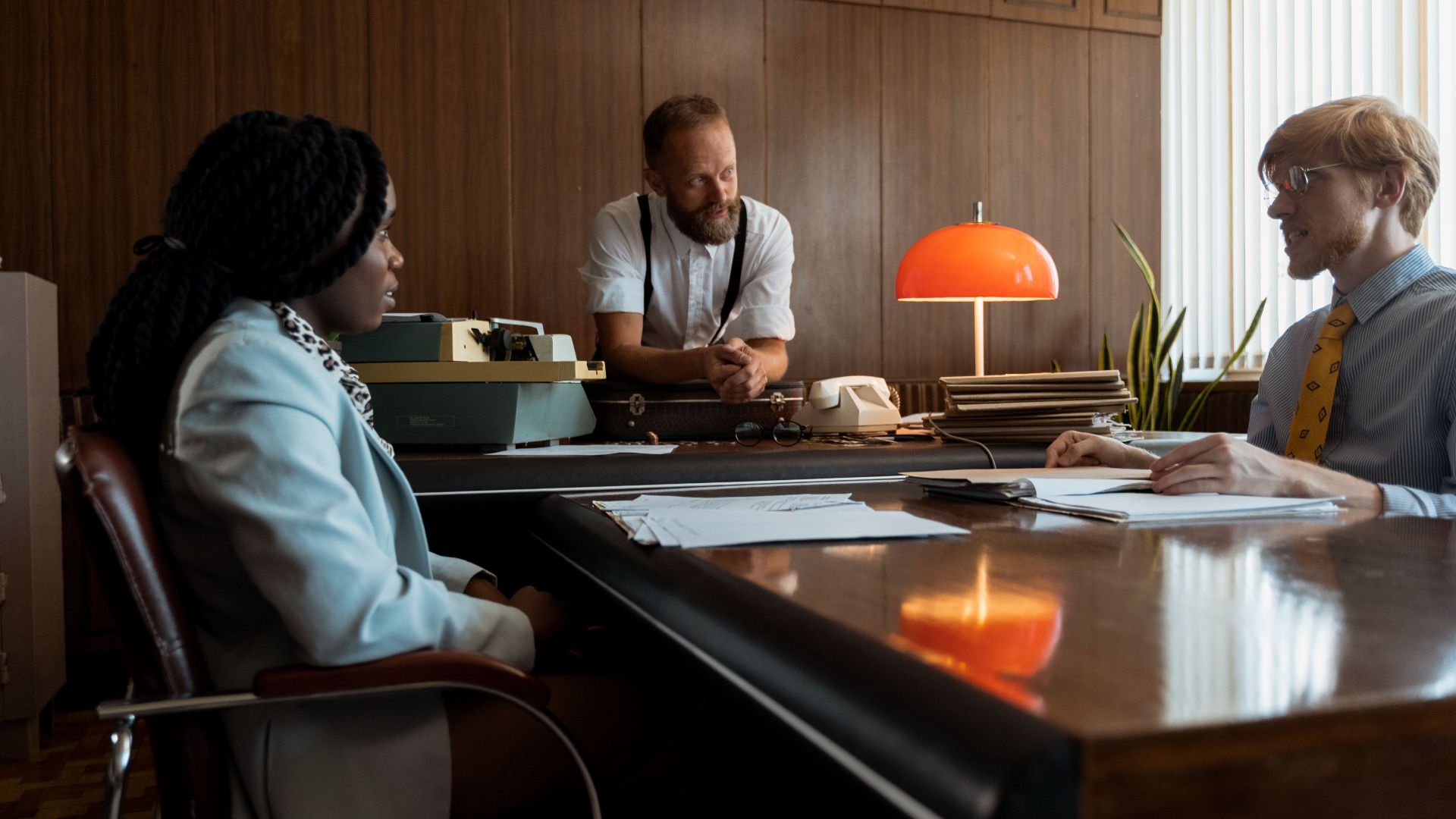 Three colleagues engage in a discussion around a vintage desk in a retro office space.