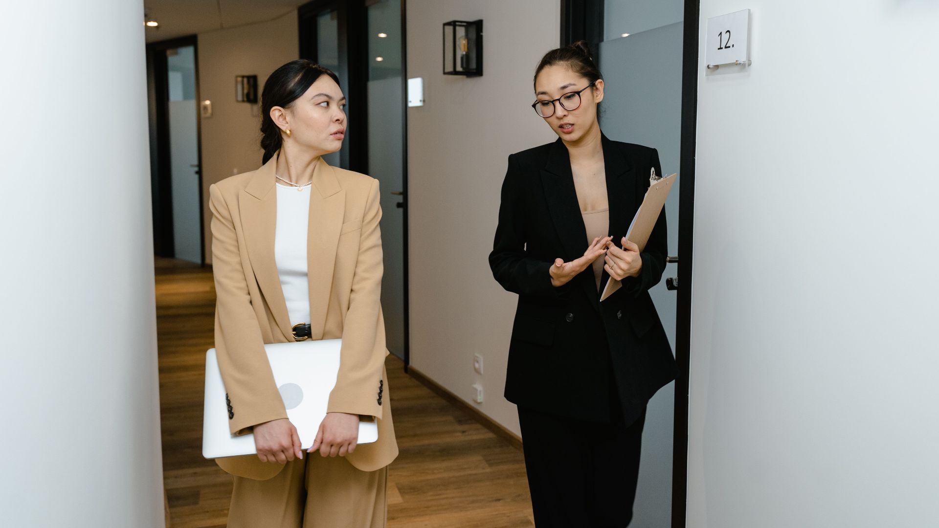 Two professional women engaged in work discussion in an office hallway, one holding a laptop.