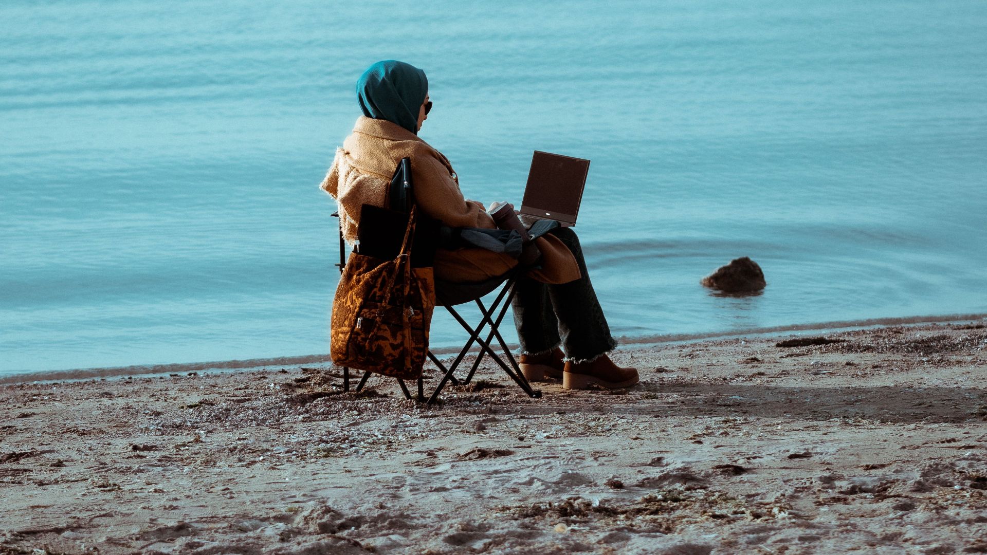 A person sitting on a chair by the water's edge, using a laptop on a serene beach.