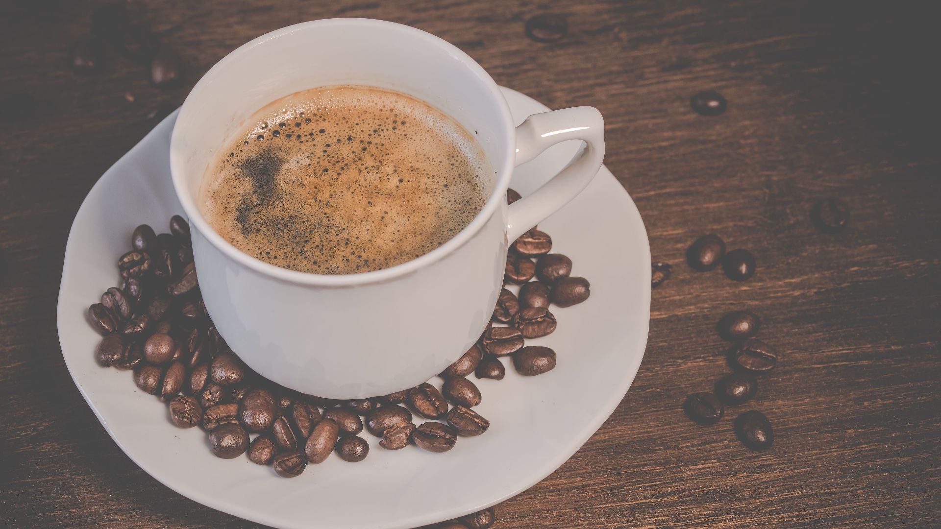A steaming espresso served in a white cup surrounded by roasted coffee beans on a wooden table.