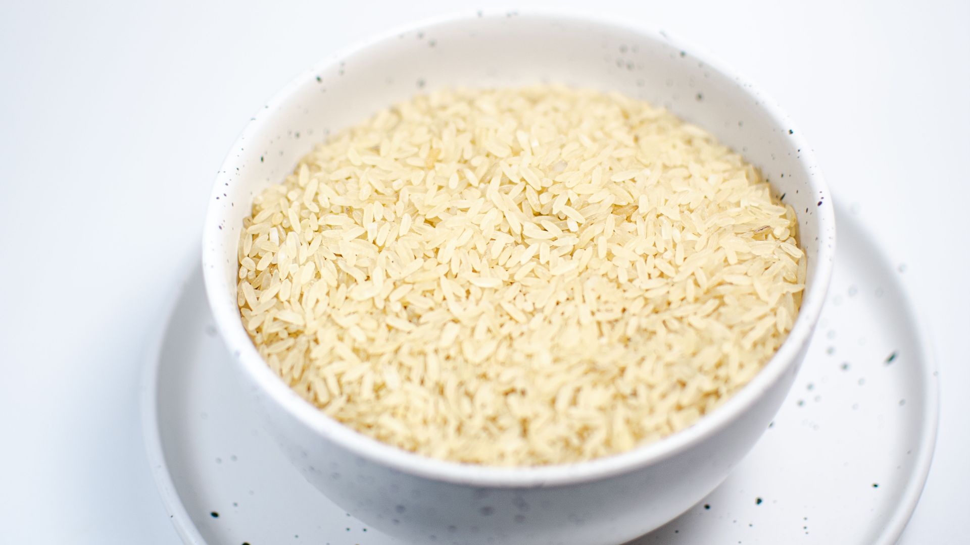 A close-up shot of uncooked rice grains in a speckled white bowl on a matching plate.