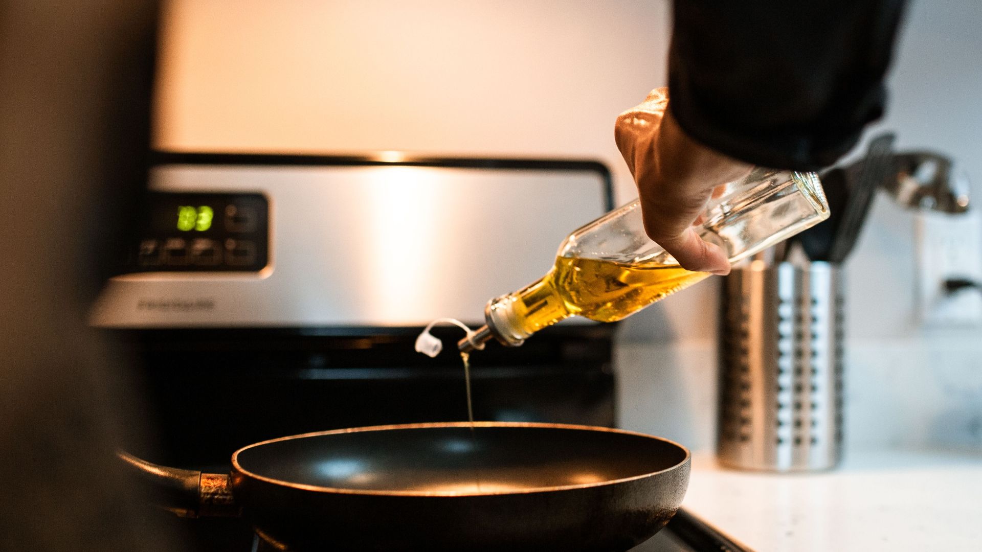 Back view crop unrecognizable person pouring olive or sunflower oil into frying pan placed on stove in domestic kitchen