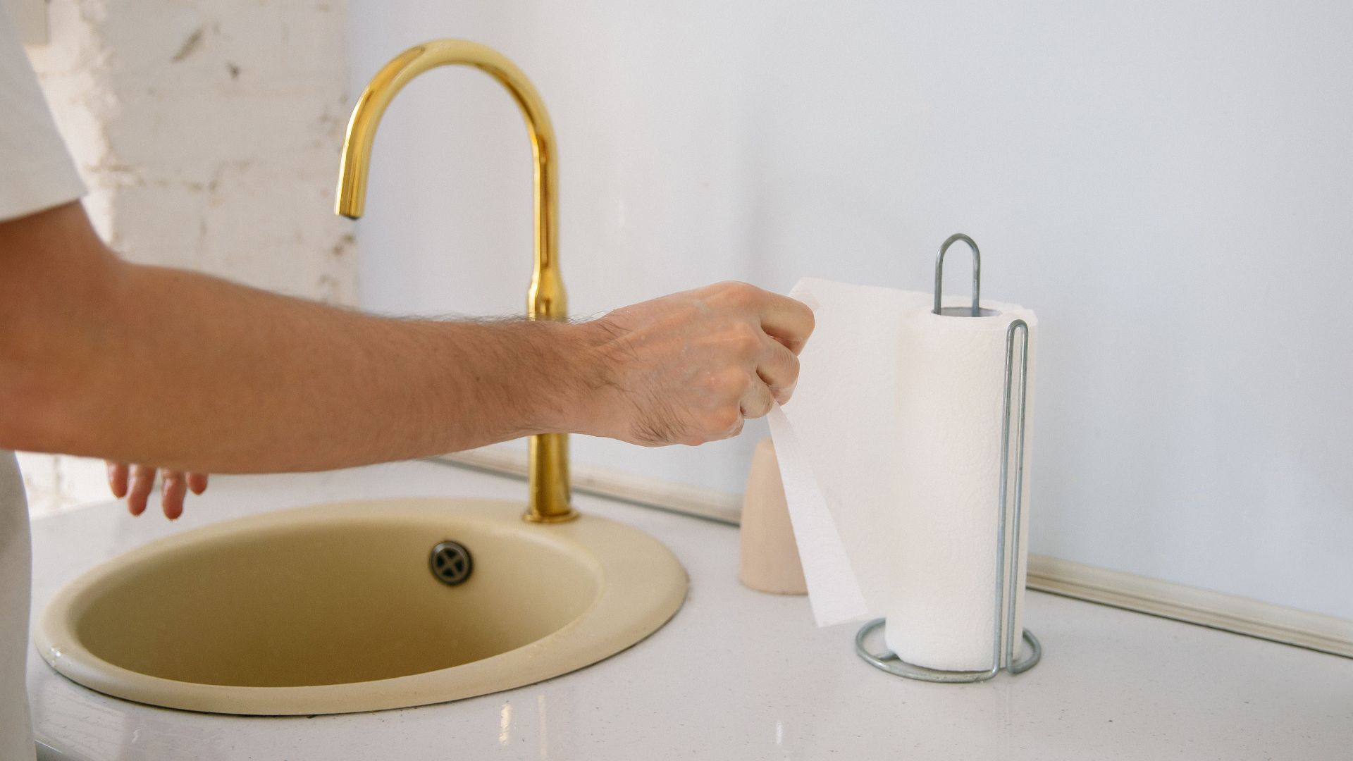 A bathroom sink with a golden faucet and hand reaching for a paper towel in a clean setting.
