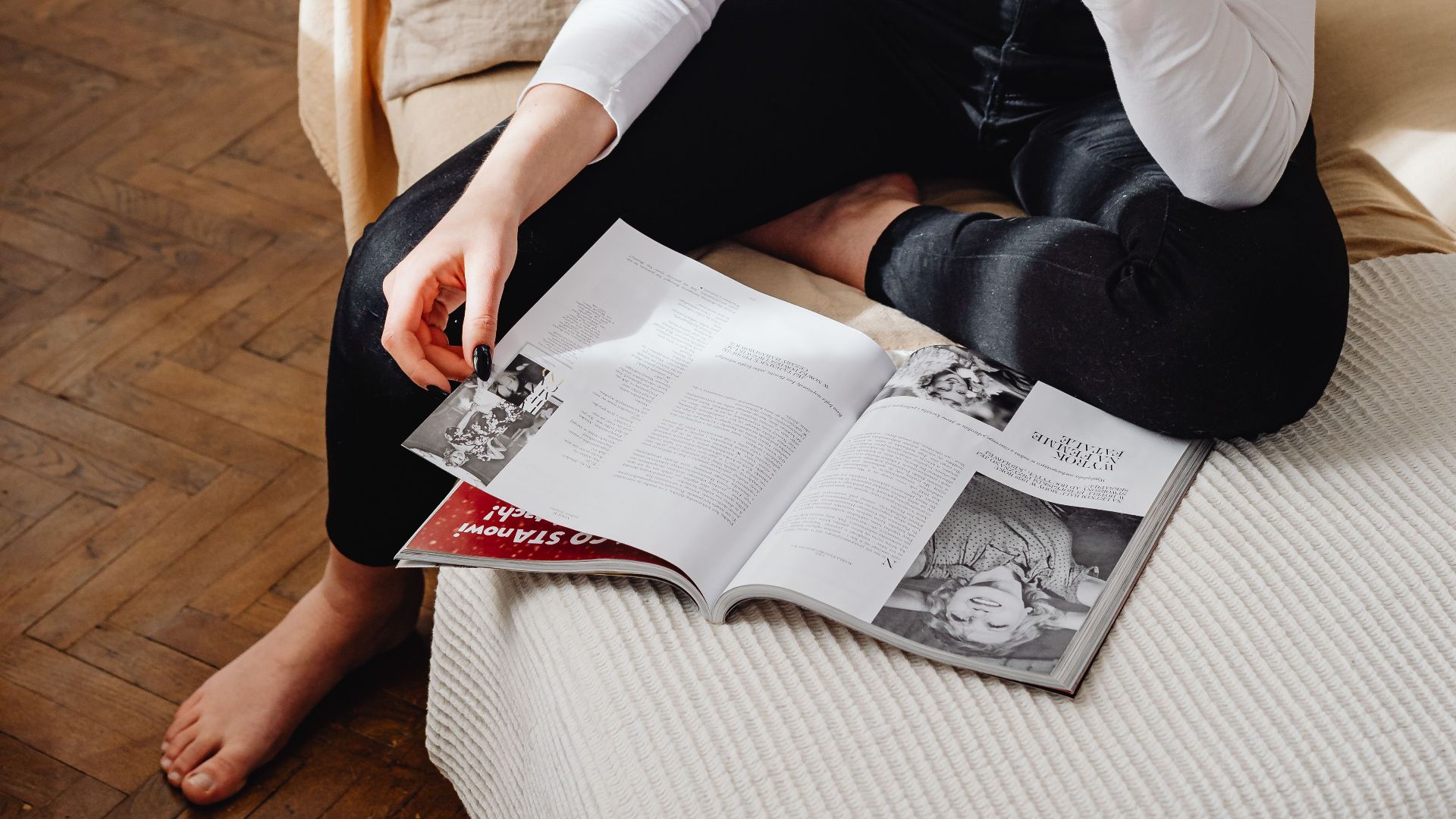 A young woman sits thoughtfully on a sofa, reading a magazine in a well-lit living room.