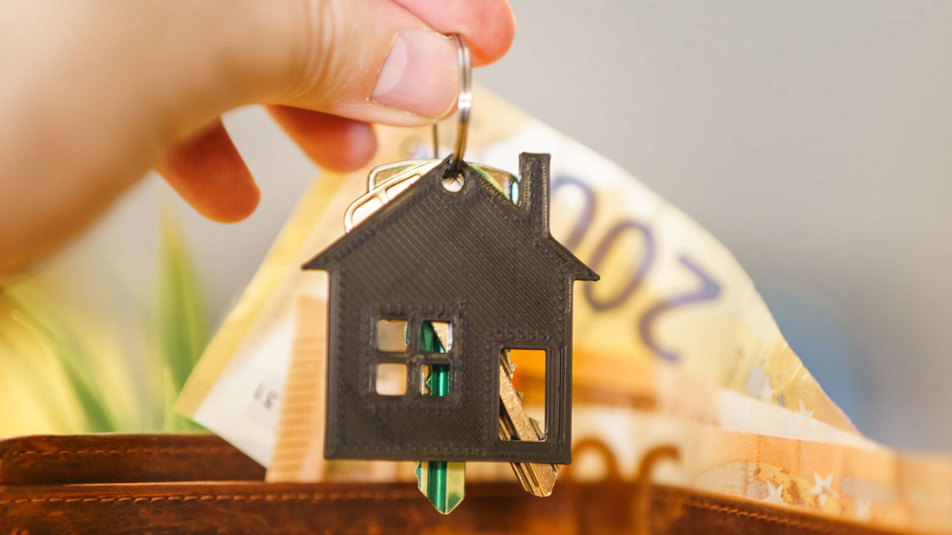 A hand holds a house-shaped keychain above an open wallet with Euro banknotes.