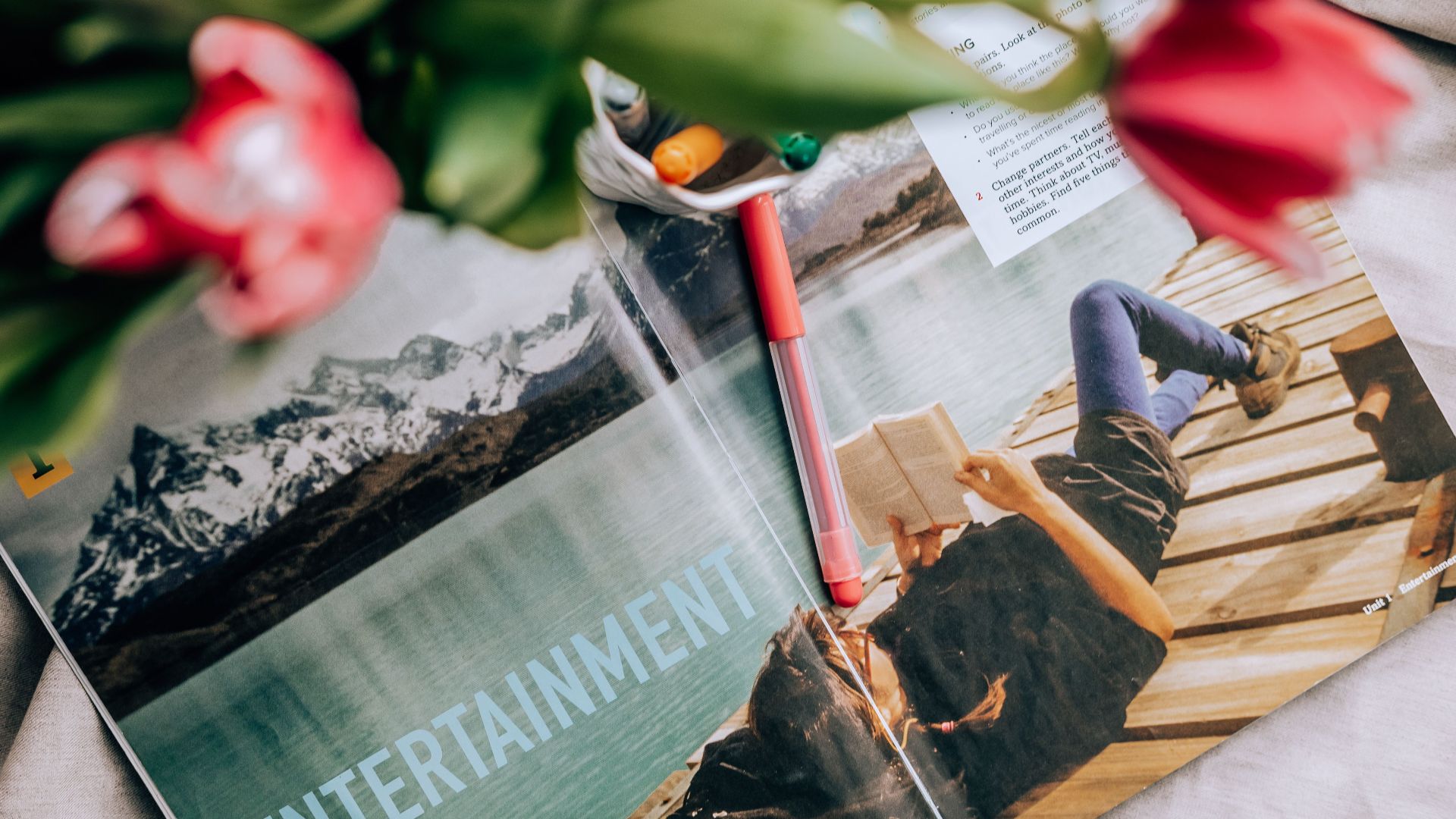Person reading on a dock by a lake with a scenic mountain view in a magazine.