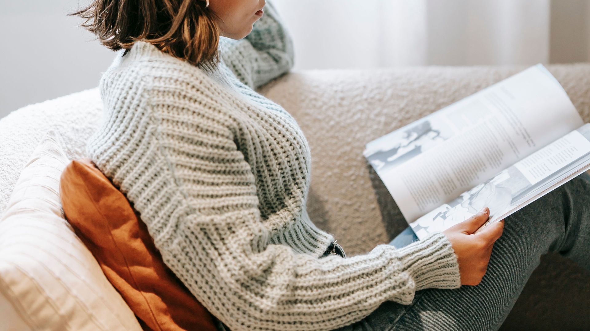 Side view of focused Asian female sitting on sofa and reading book while spending leisure at home