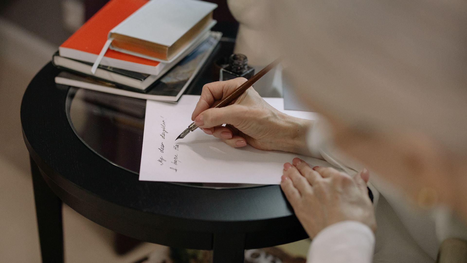 A person writes on white paper using a fountain pen, surrounded by books on a table.
