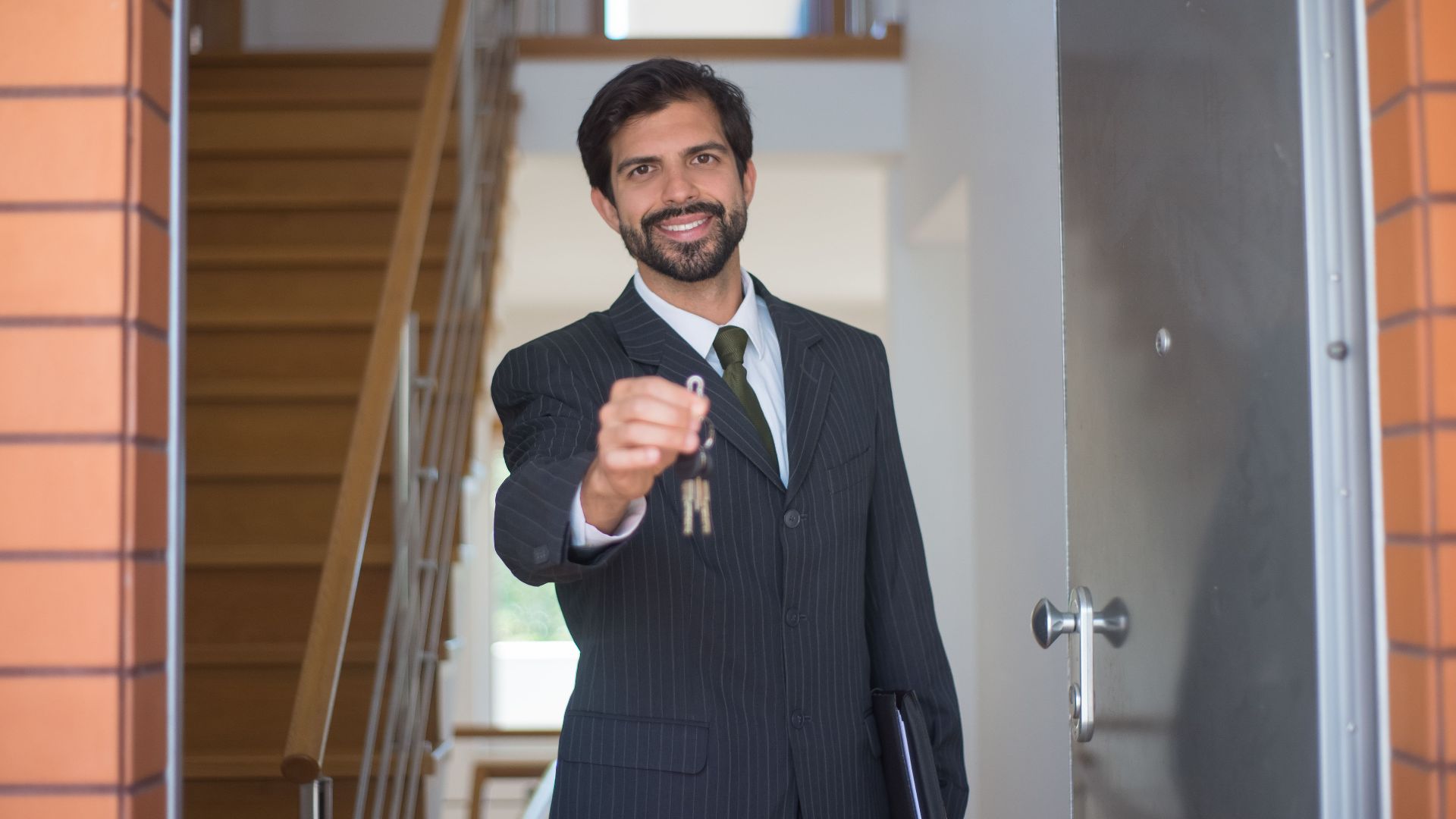 Smiling realtor in a black suit handing keys through an open door inside a modern house.