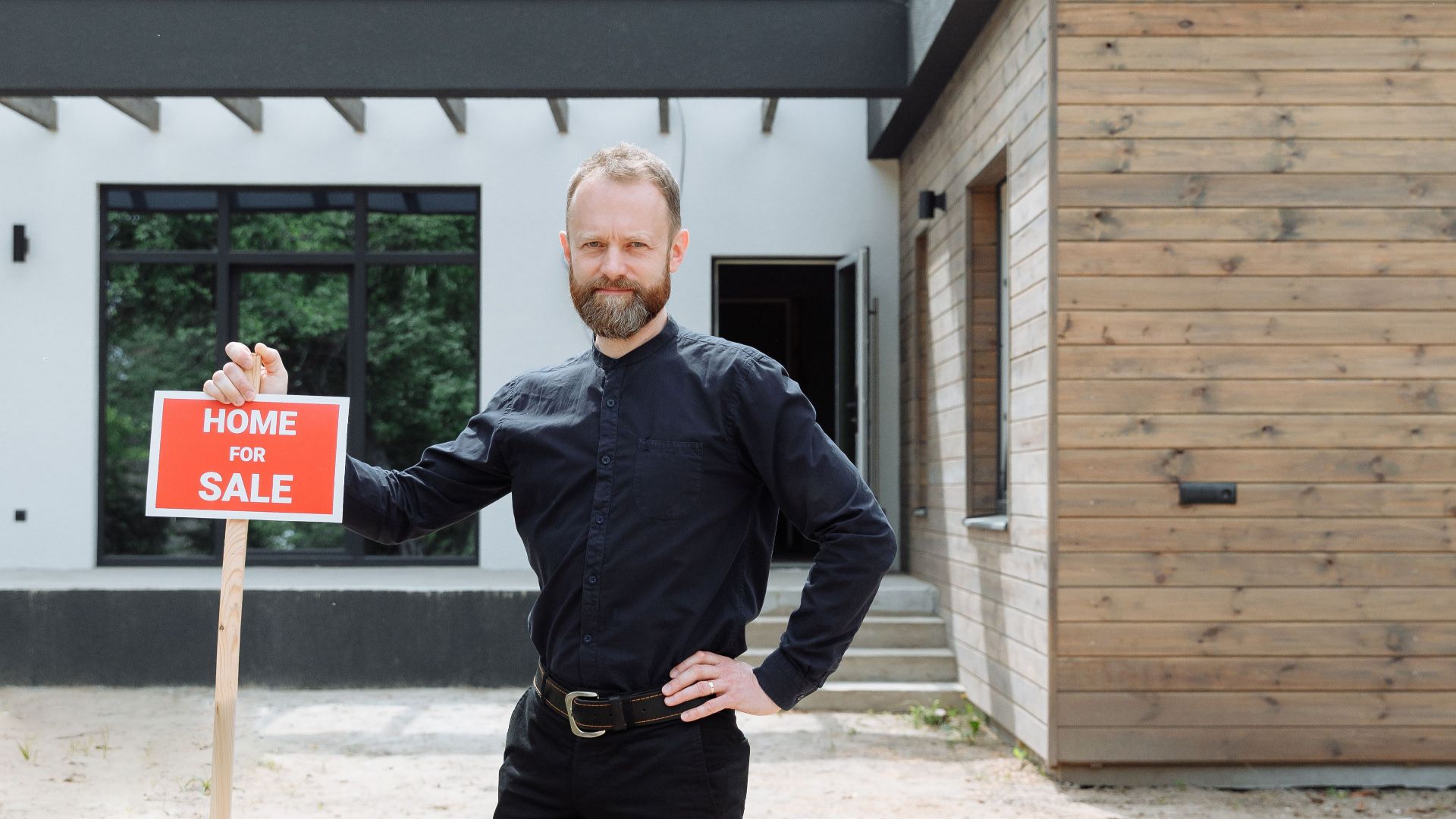 A real estate agent stands outdoors holding a 'Home for Sale' sign in front of a modern house.