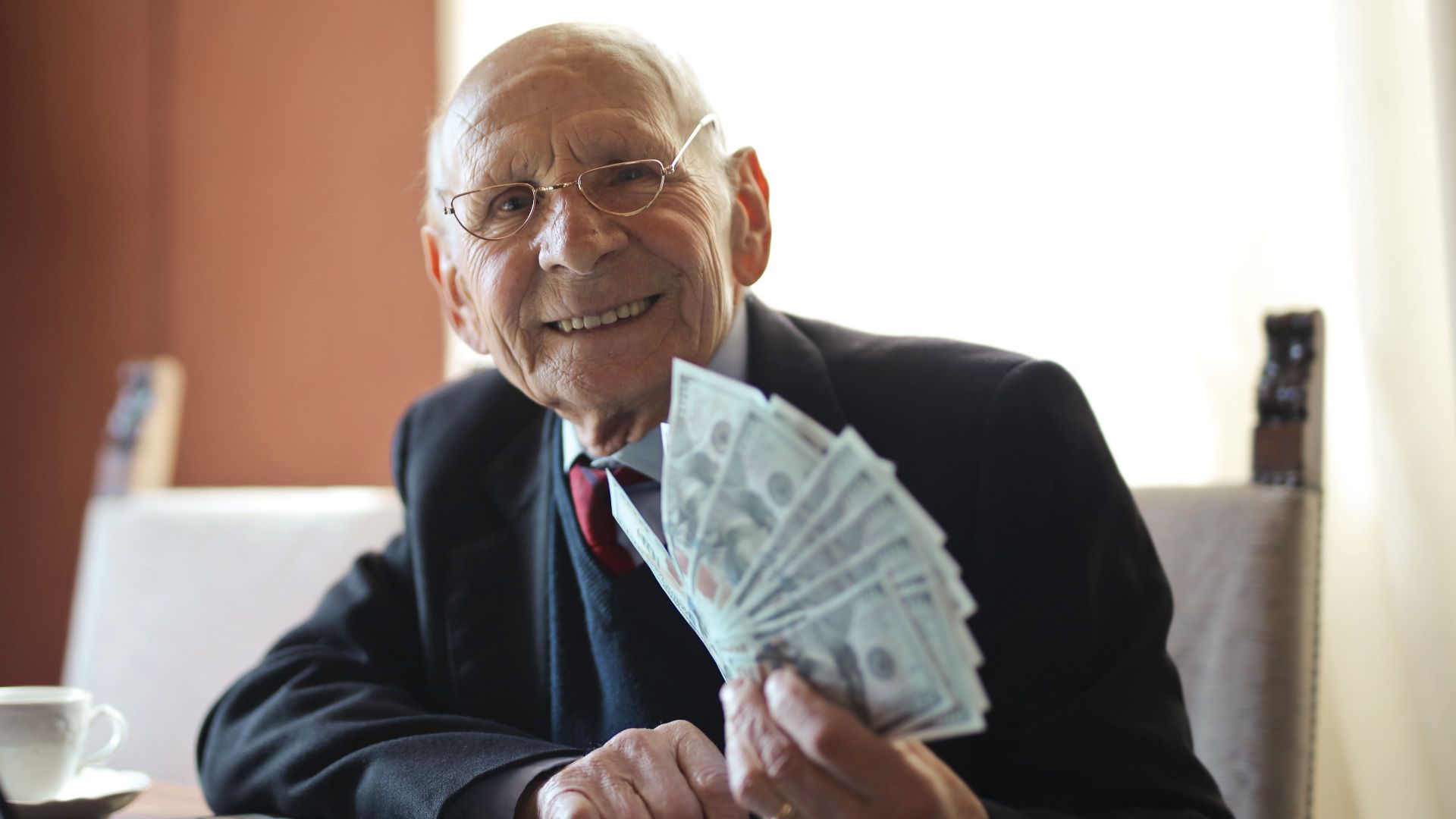 Positive senior businessman in formal suit and eyeglasses holding bunch of bills while working on laptop at table and looking at camera