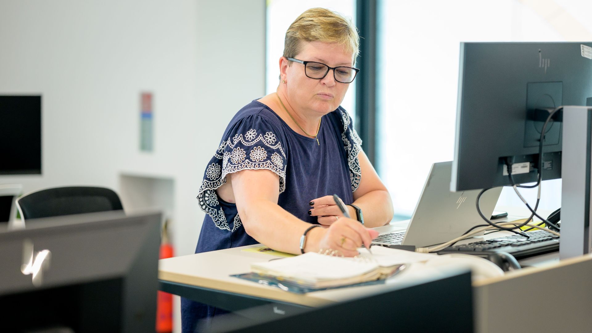 Professional woman wearing eyeglasses working at an office desk with a laptop and notebook.