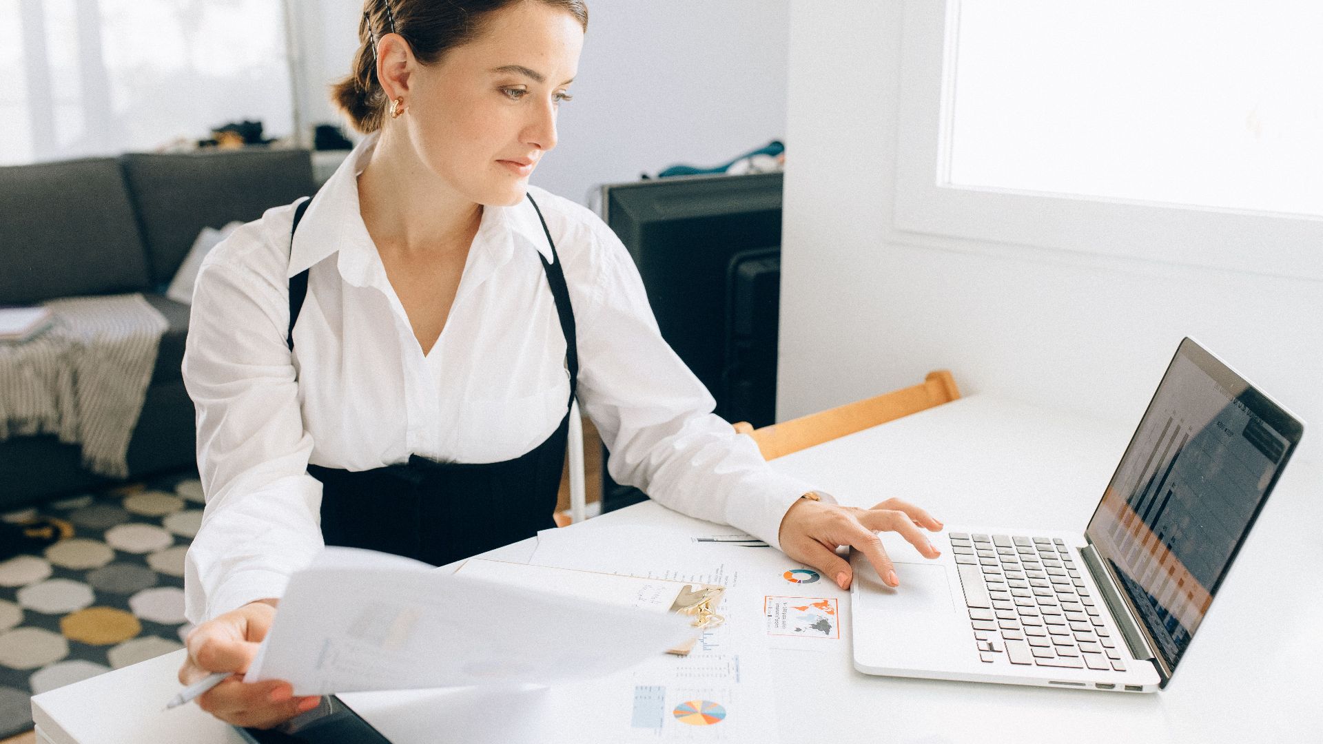 Woman at desk reviewing business reports on laptop, holding papers in a modern office setting.