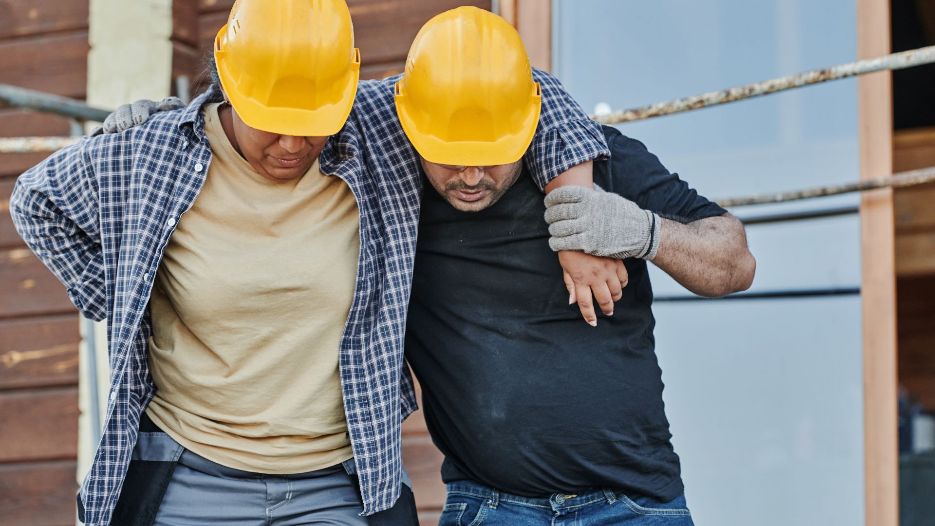 Two construction workers in hard hats assisting each other at a building site, highlighting teamwork.