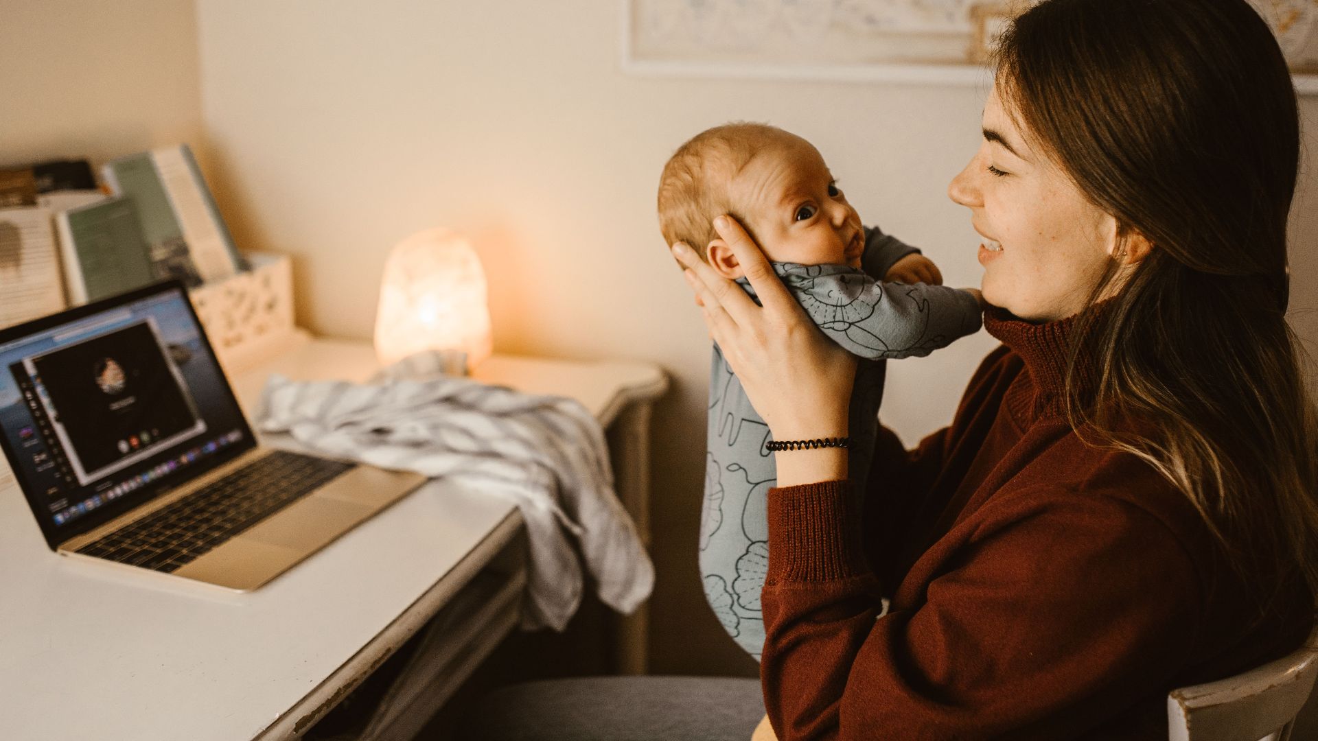 Mother holding baby while video calling from home office. Cozy and heartwarming indoor scene.