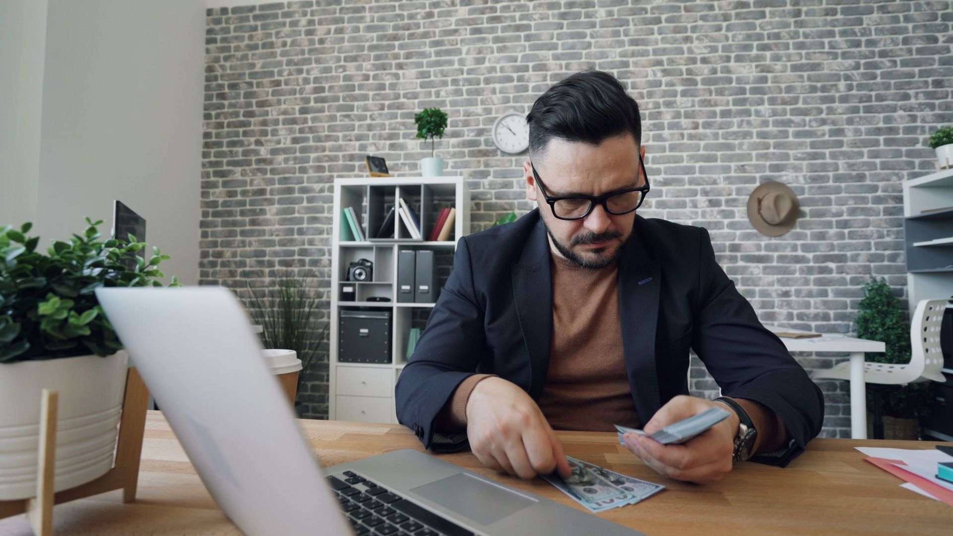 Businessman counting cash at a modern office desk with a laptop, showcasing finance management.