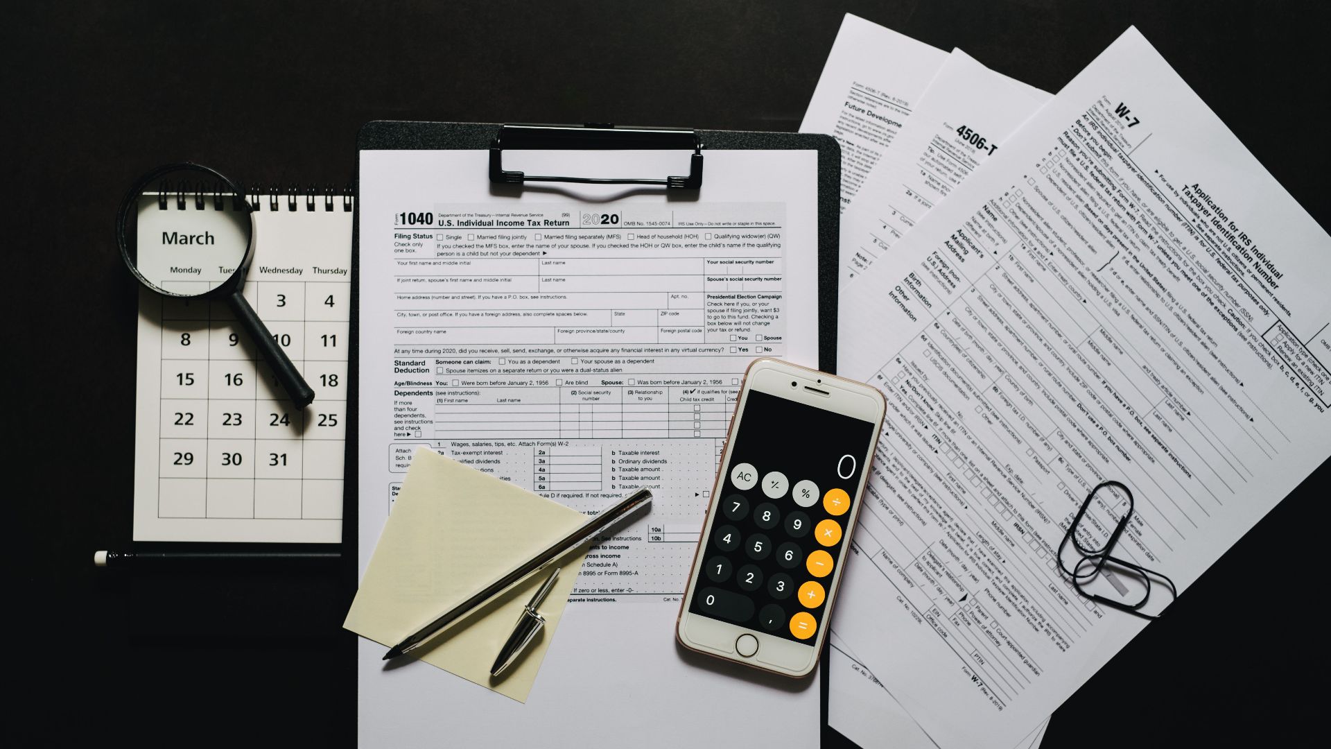 Top view of tax documents, calculator, magnifying glass, and calendar on a black surface.