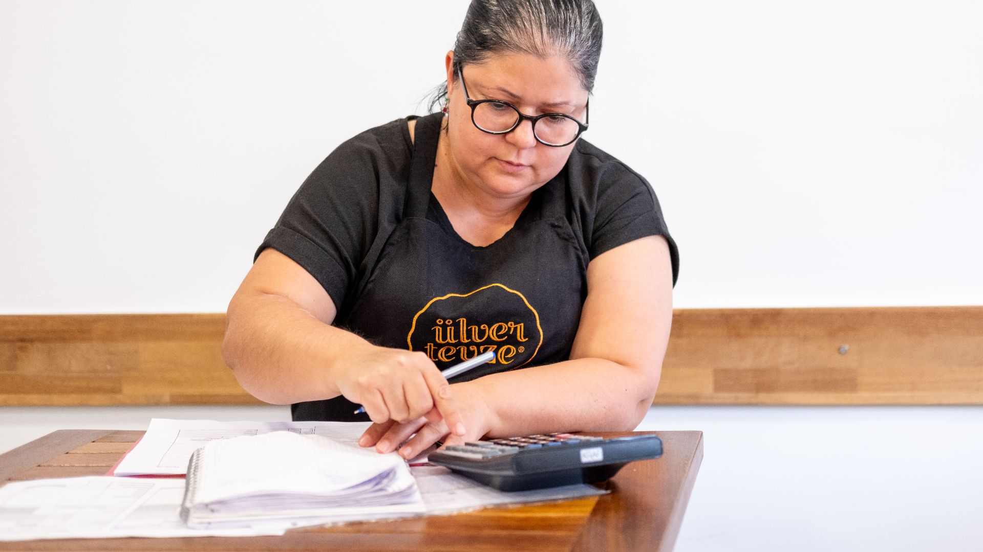 Middle-aged woman using a calculator while working on financial tasks at a wooden desk indoors.