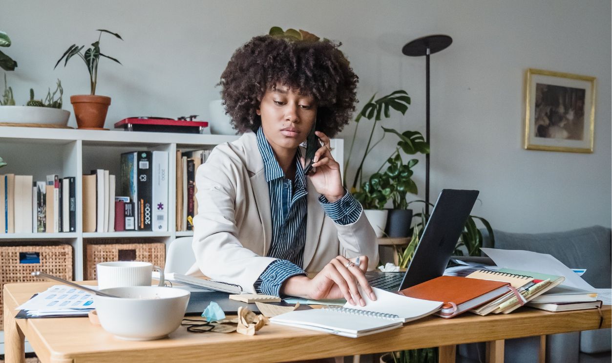 Woman working from home office, multitasking with laptop and phone, surrounded by documents.