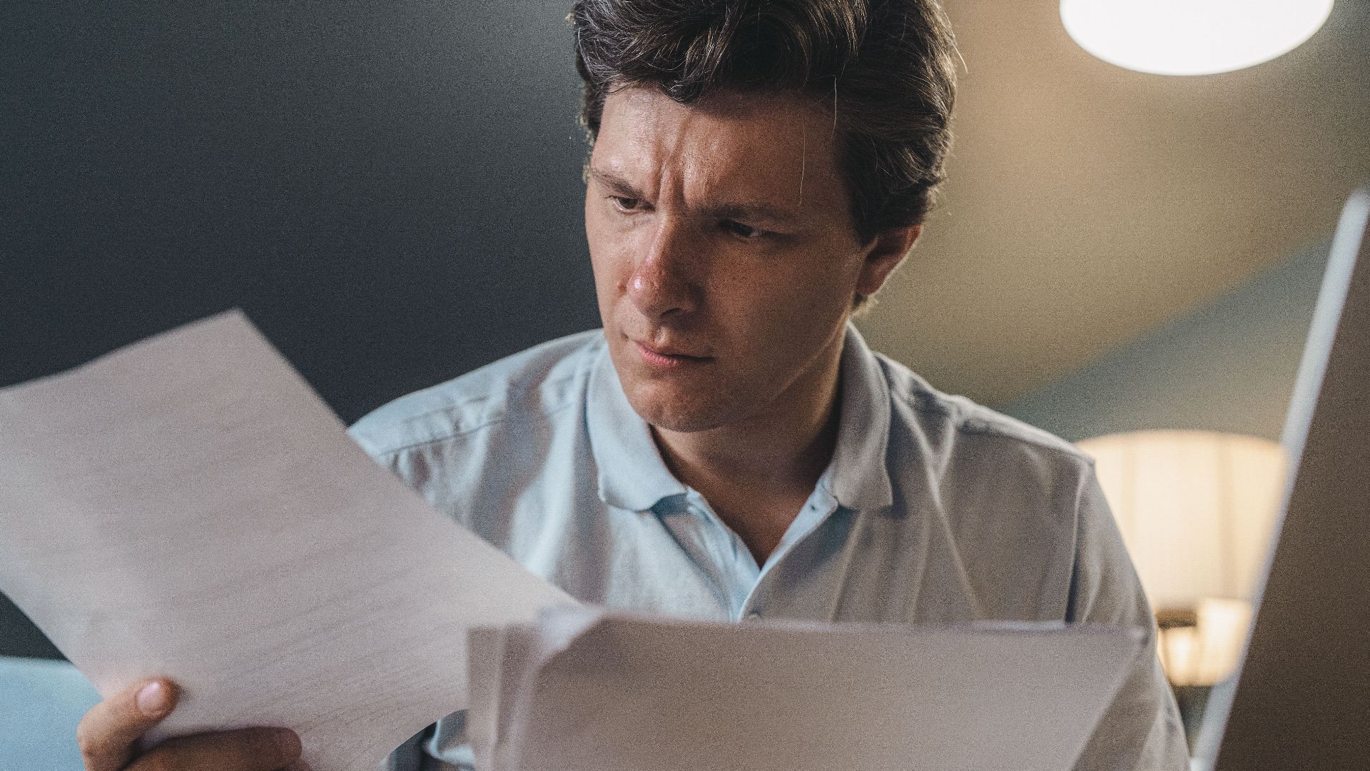 A man intently examines papers, seated indoors under warm lighting, focusing on his work.