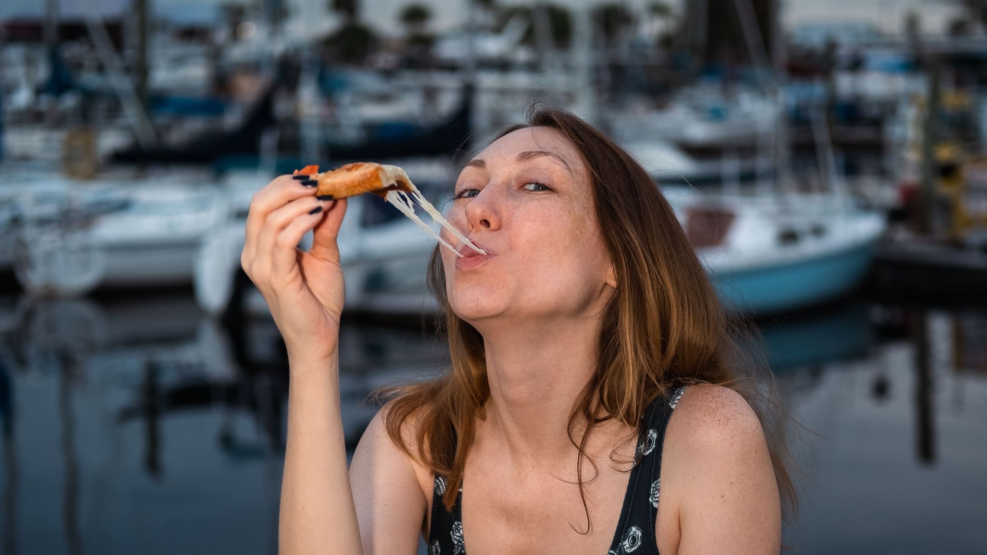 A woman enjoys a slice of pizza by the marina at twilight, capturing a relaxed lifestyle moment.