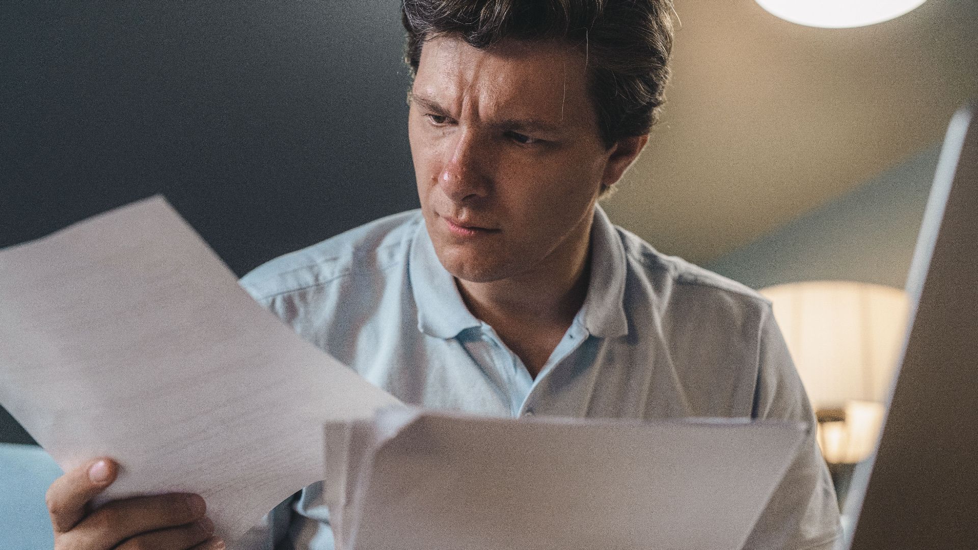 A man intently examines papers, seated indoors under warm lighting, focusing on his work.