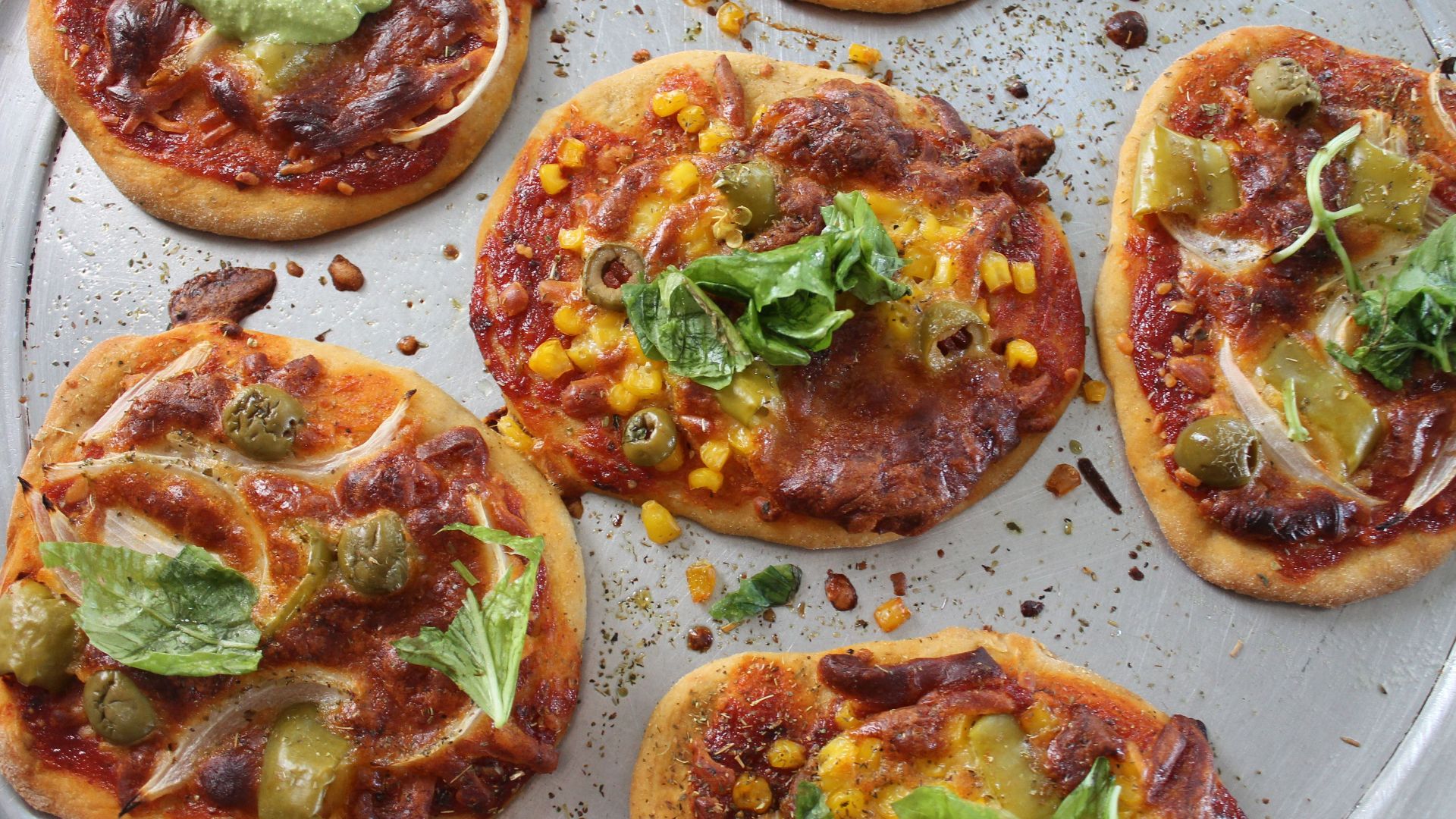 Close-up of assorted pizzettas with toppings like olives and basil on a baking tray, emphasizing their deliciousness.
