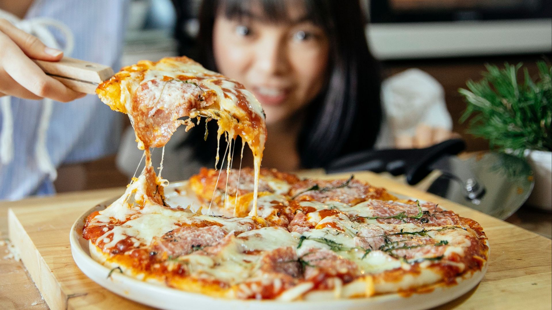 Unrecognizable women sharing slice of delicious pizza with melted cheese and salami from big plate using wooden scapula