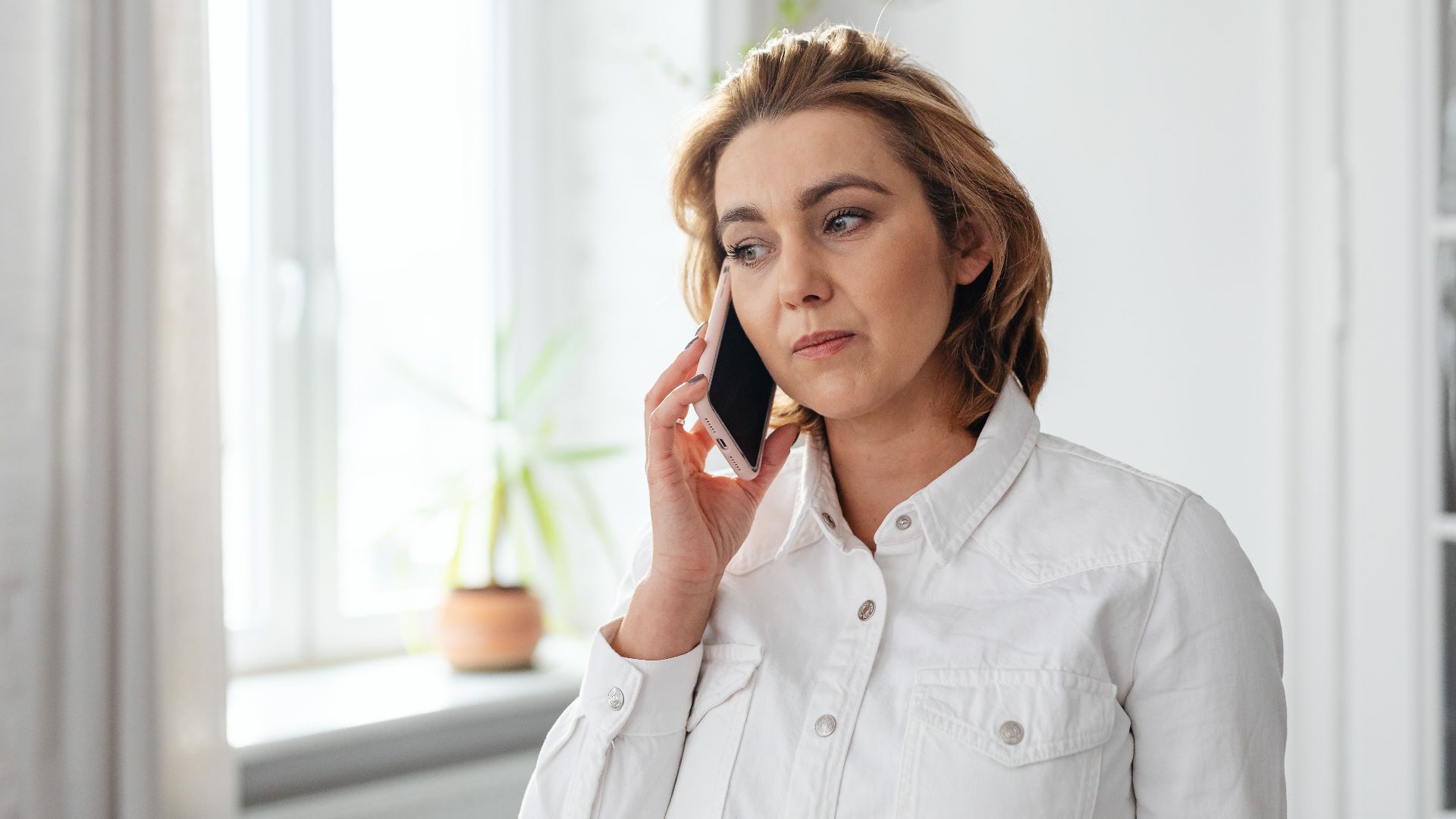 Adult woman talking on a smartphone by a window in a bright room.