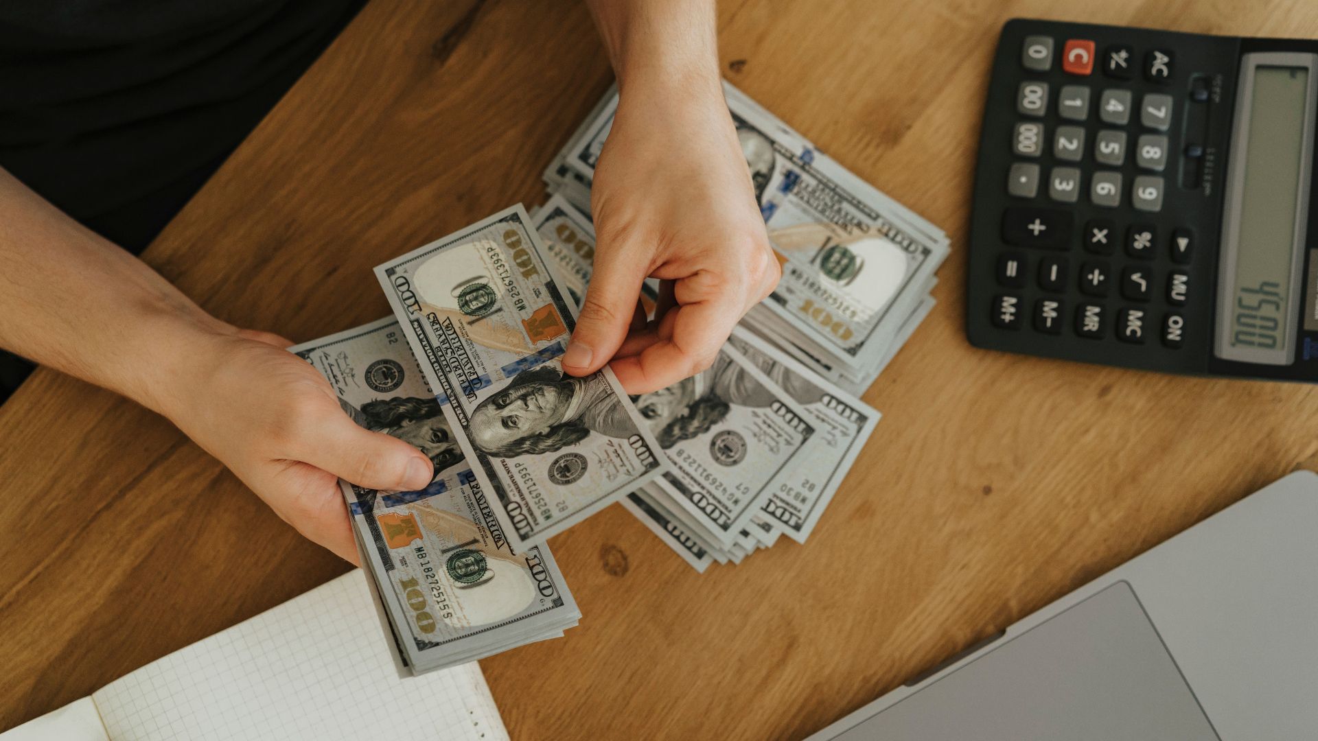 Close-up of hands counting dollar bills with calculator, notebook, and laptop on wooden table.