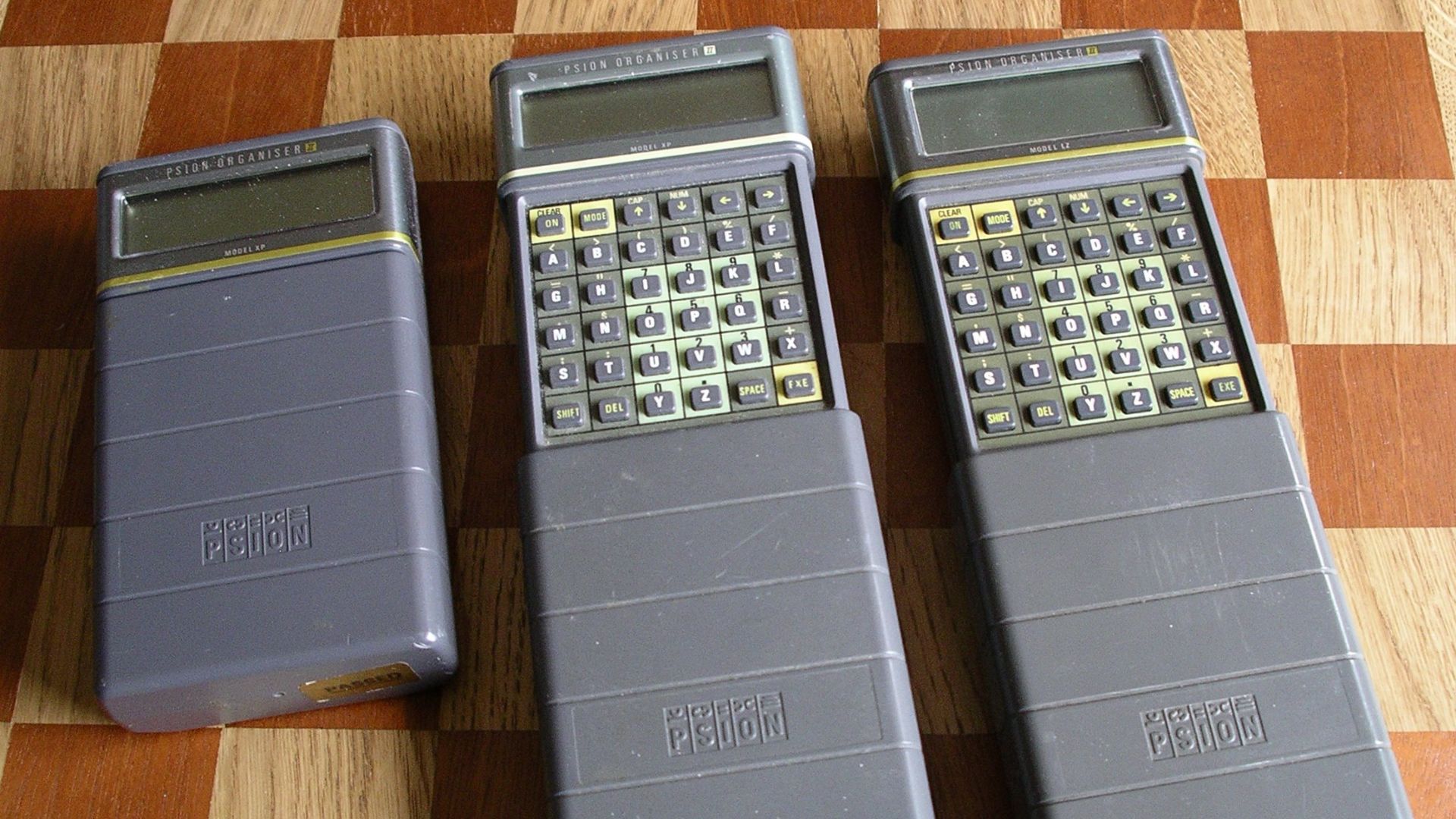 Picture shows one closed Psion Organiser and two Psion Organisers with the protective cover open. Model XP and Model LZ are shown. The background shows 5 cm squares. Photo taken by me on 17 October 2006.