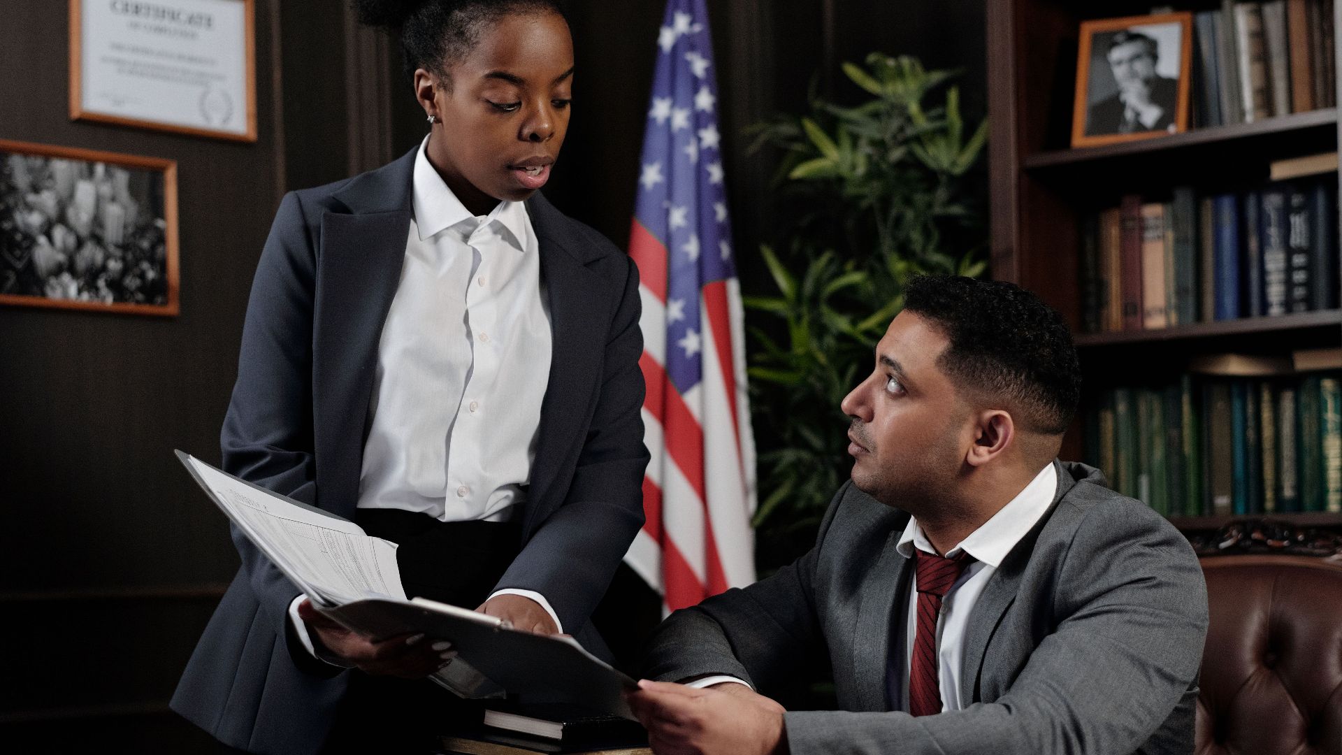 Two professionals discussing documents in an office with an American flag in the background.