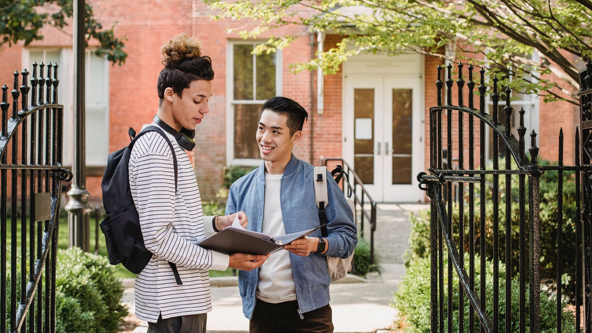 Cheerful diverse male classmates with paper document in folder standing close and discussing task at university entrance
