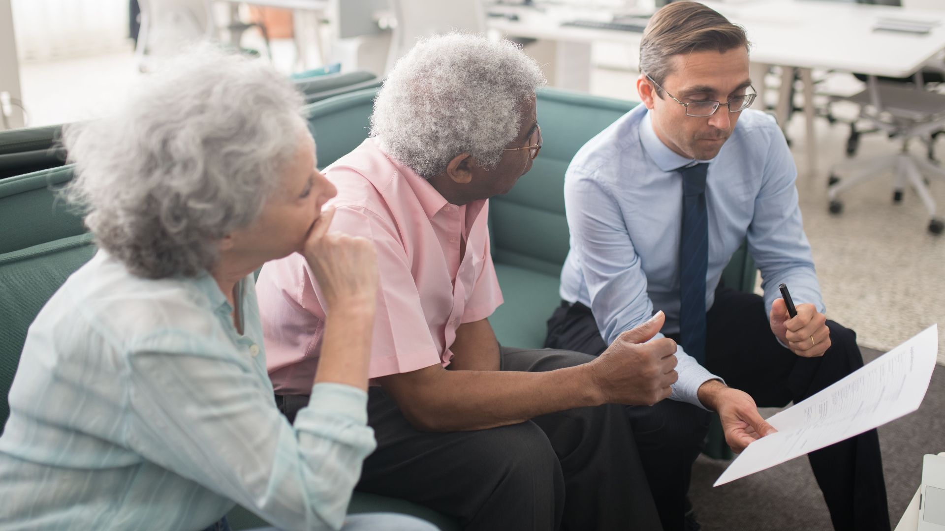 Business professional consults elderly clients in an office setting. Collaborative discussion, paperwork visible.