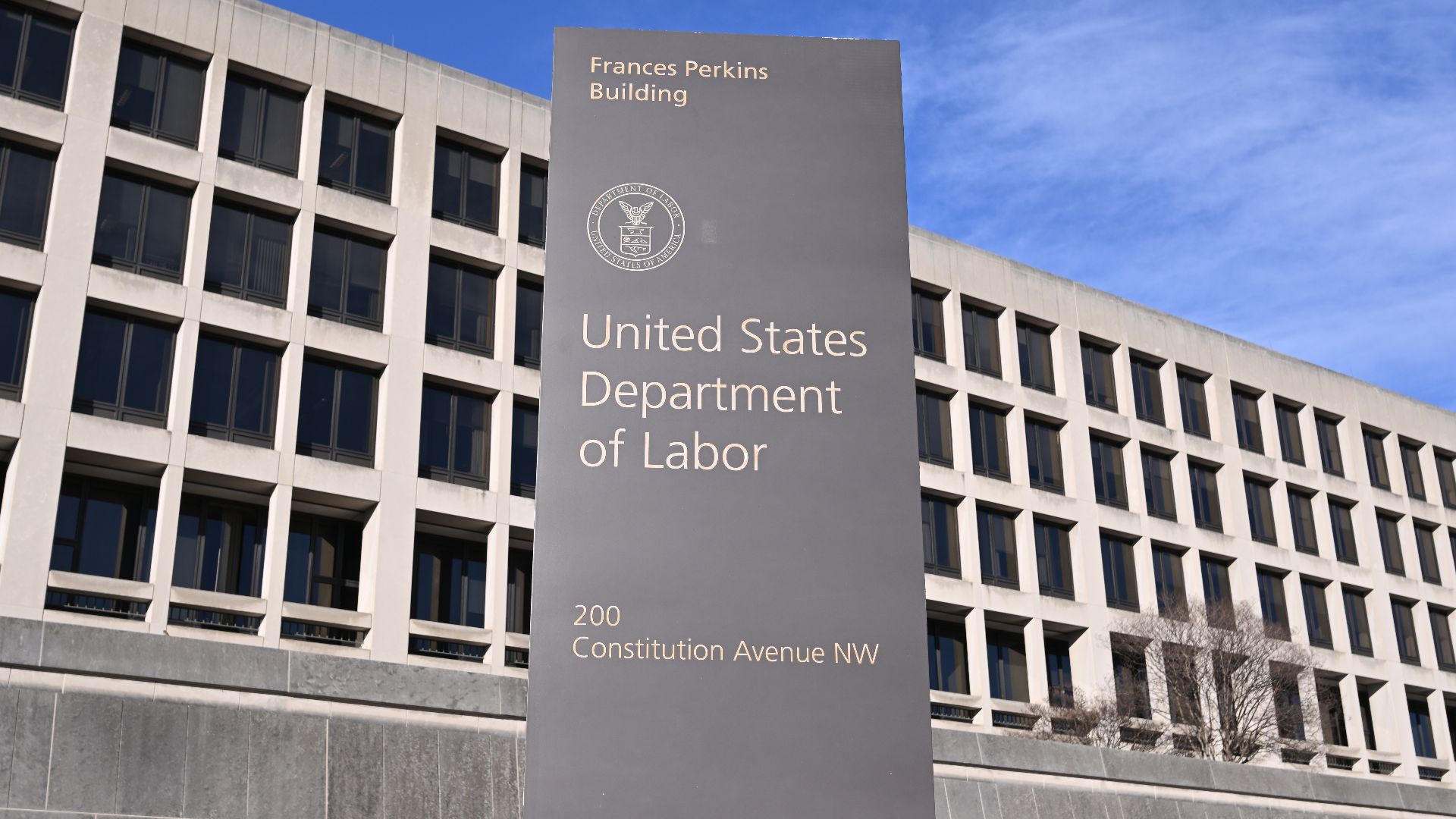 A sign on Constitution Ave for the United States Department of Labor with part of the Frances Perkins building behind it. 200 Constitution Avenue NW, Washington, DC 20004.