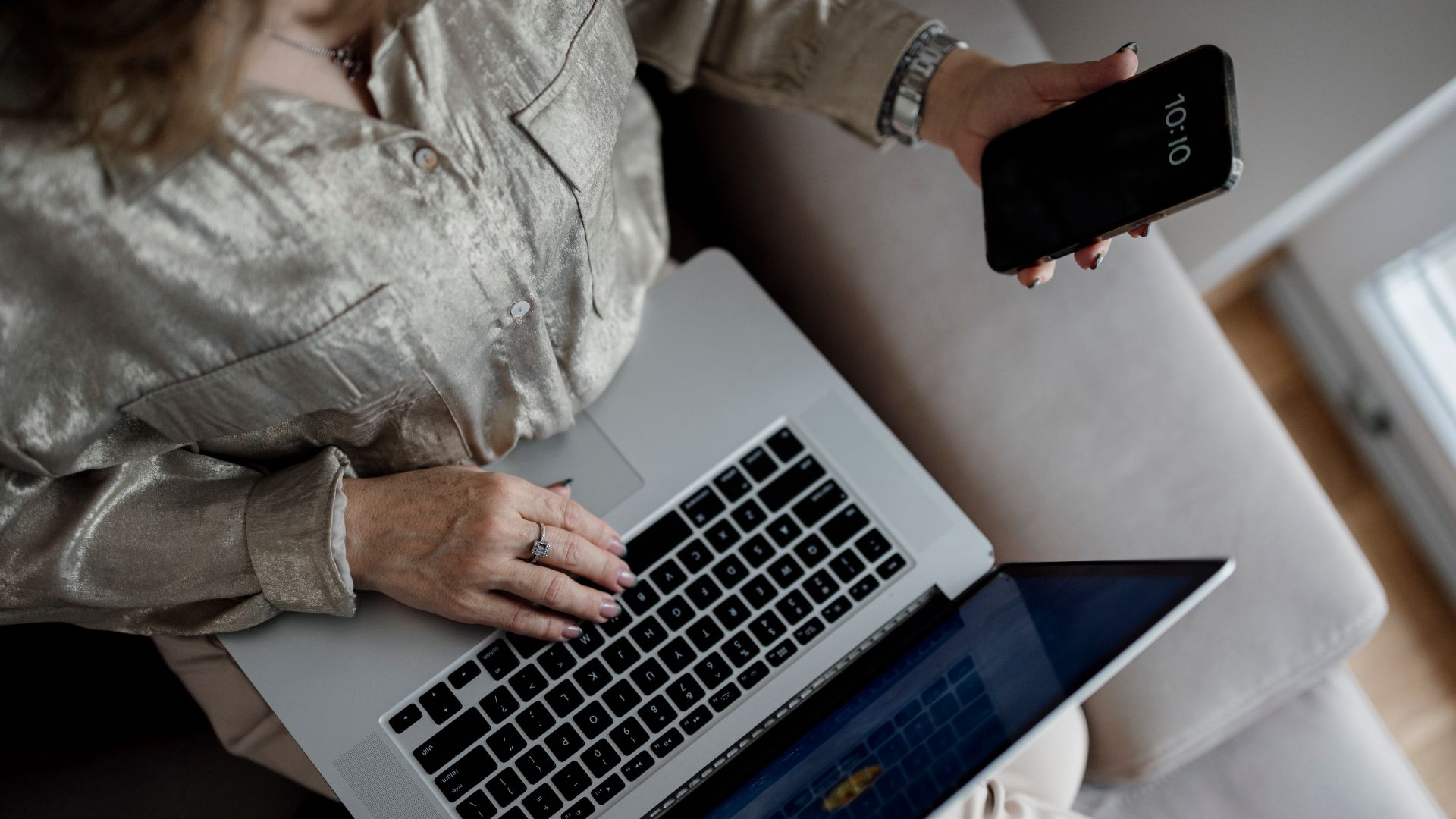 A person sits on a couch with a laptop open, typing while holding a smartphone in one hand. The space is bright with natural light coming through a window.
