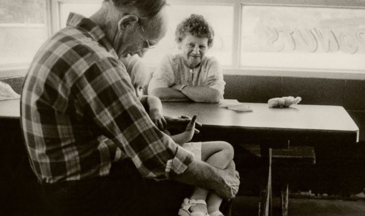 At a corner table in a diner, a man as he holds granddaughter in his lap while his wife watches, Mariposa, California, 1984.